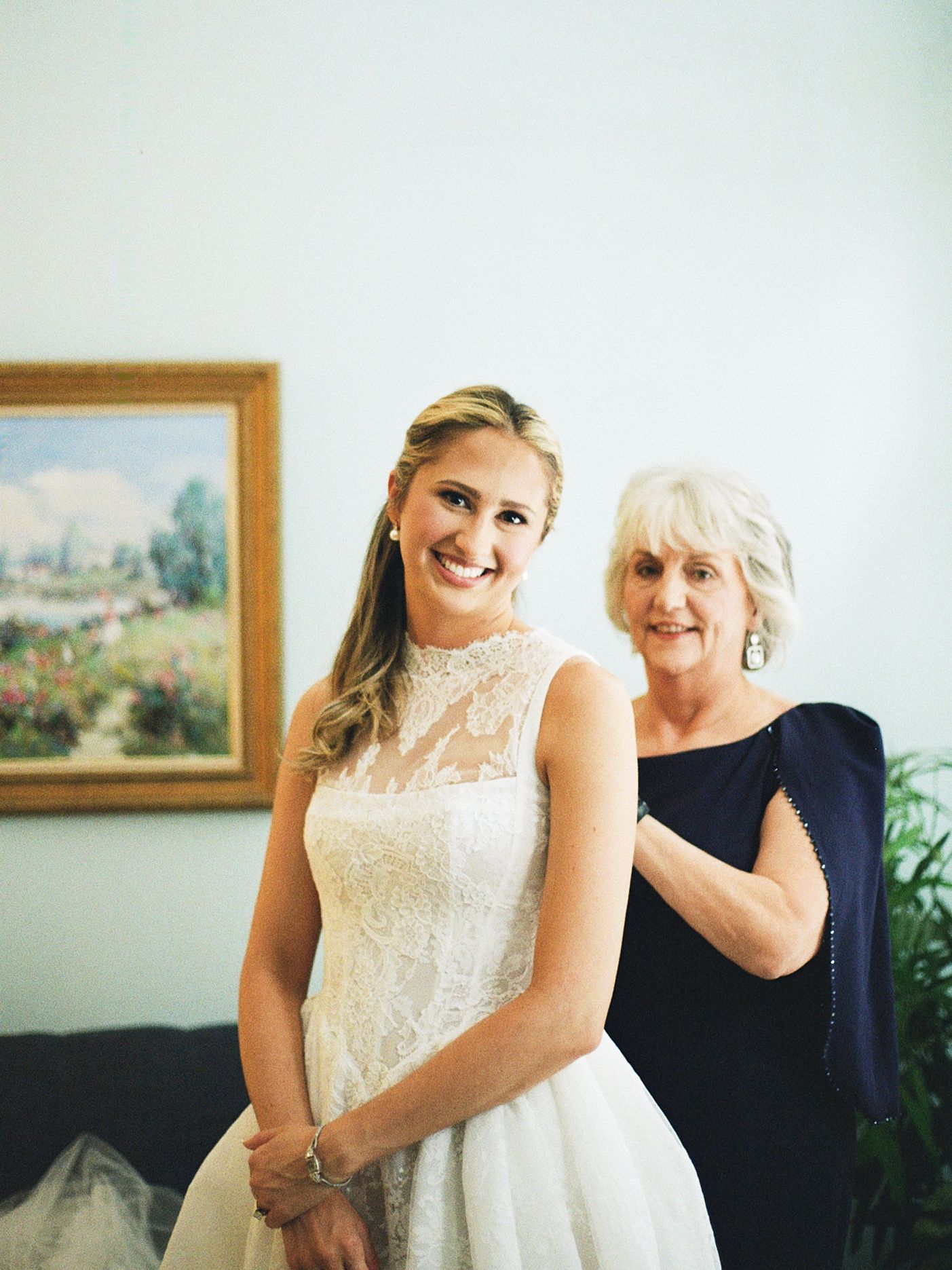 Bride in lace wedding gown smiles with older woman in navy dress helping with final preparations before ceremony.
