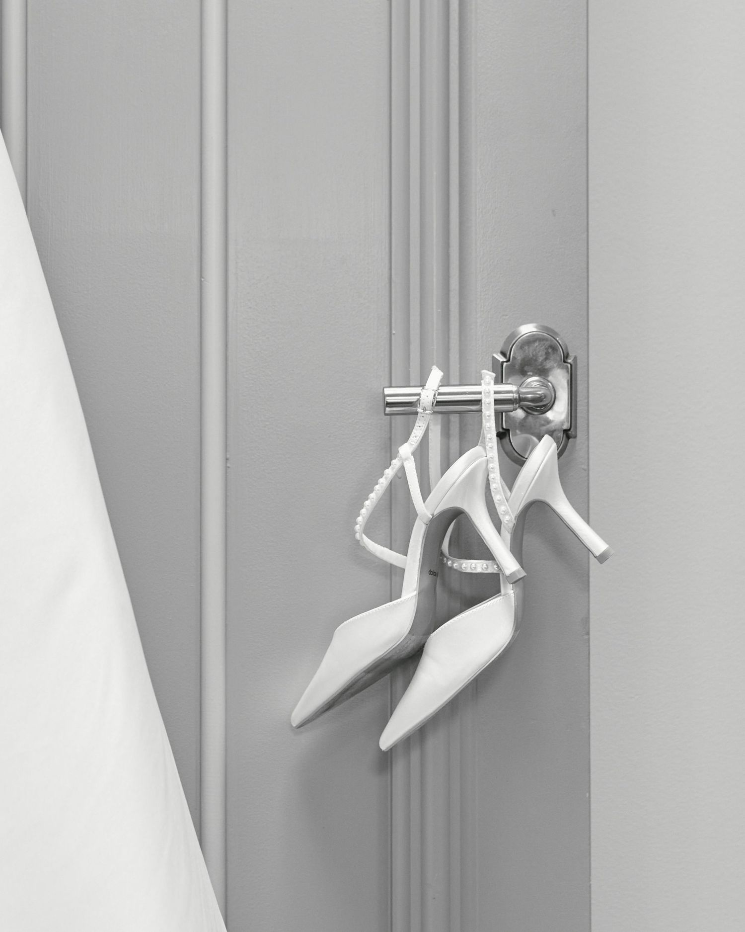 Elegant white bridal heels hanging on a door handle in a black and white photo.