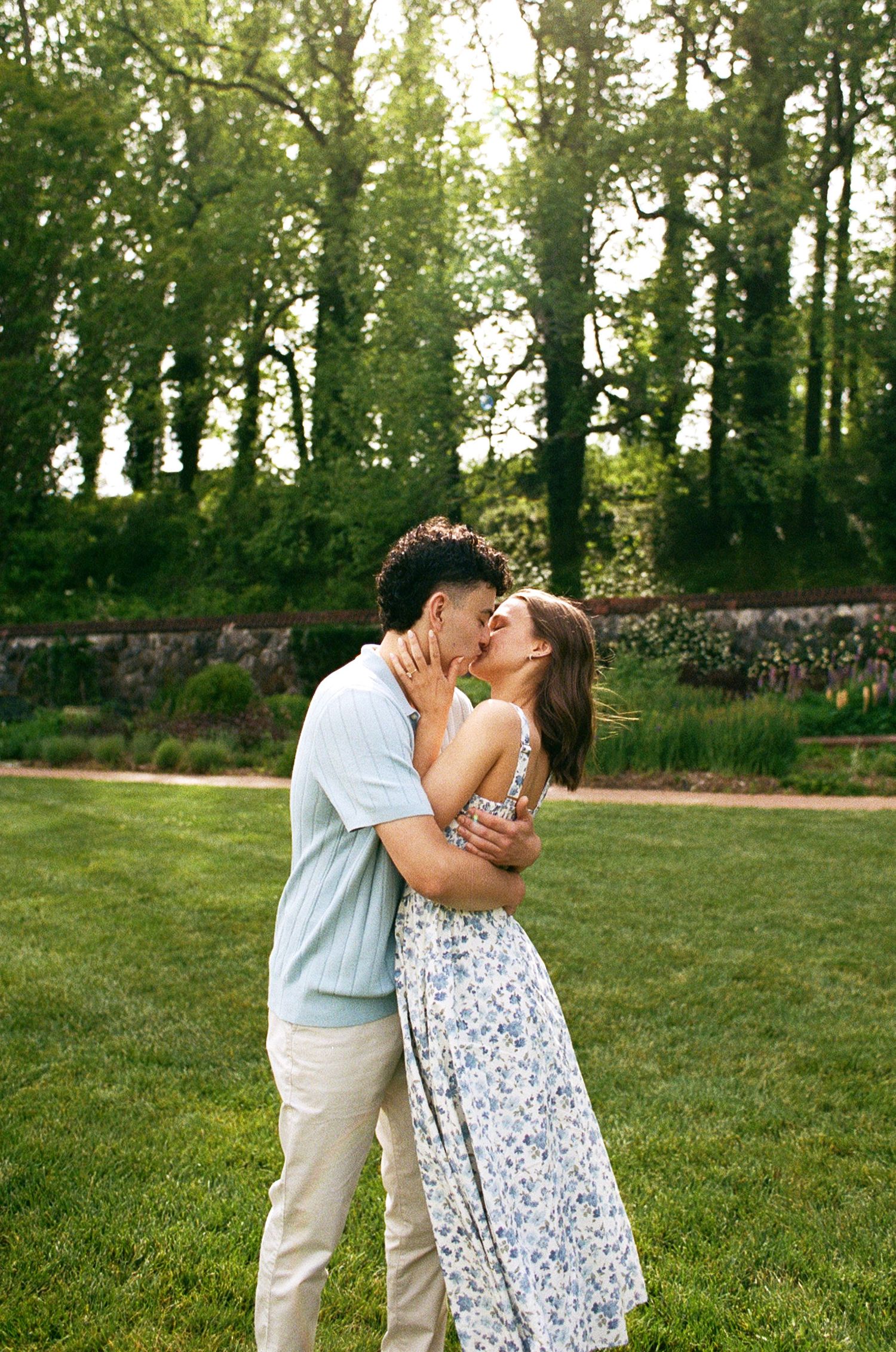 A couple shares a romantic kiss while embracing on a lush green lawn surrounded by tall trees.