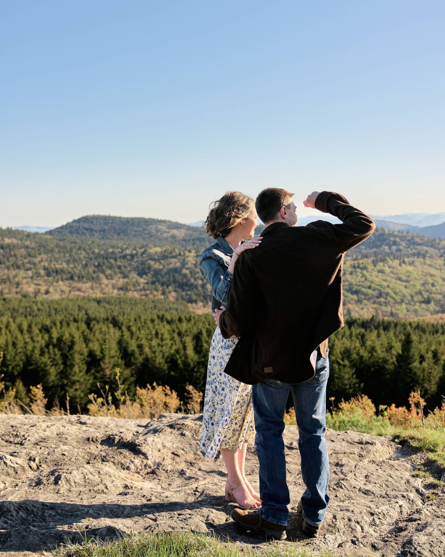 Couple standing on rocky hilltop overlooking vast forested mountain landscape under clear blue sky.