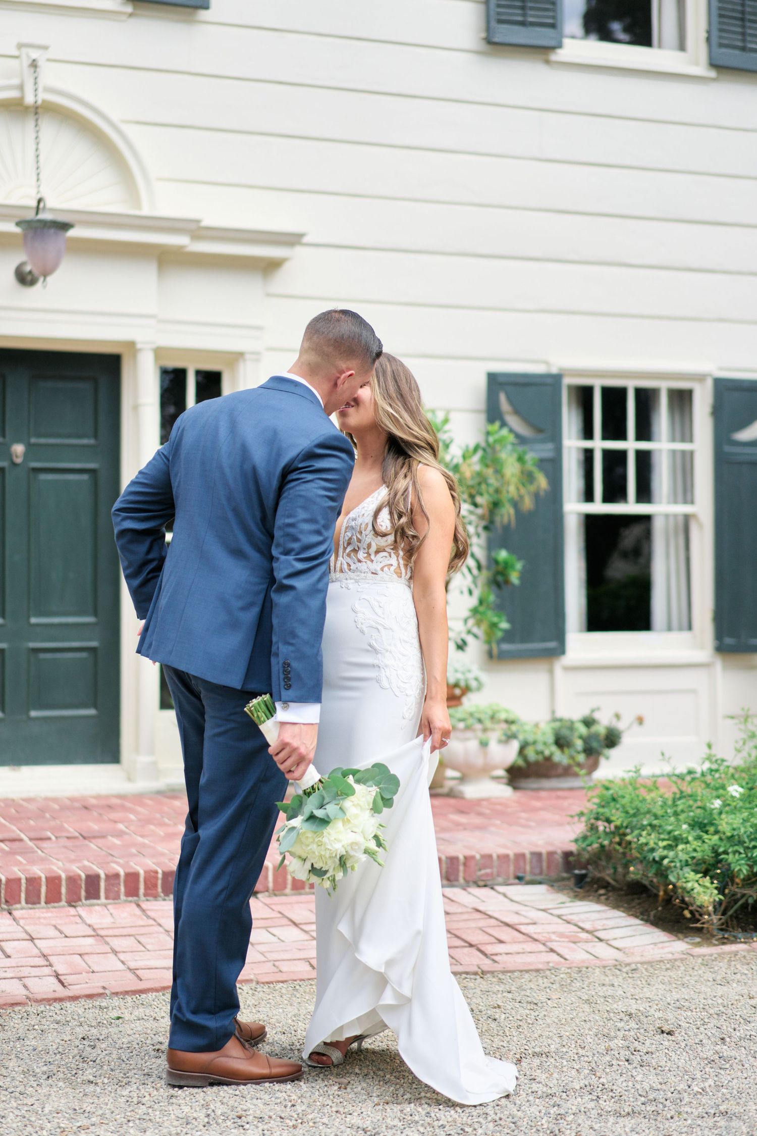 Wedding couple pose intimately by white building with green door and brick pathway.