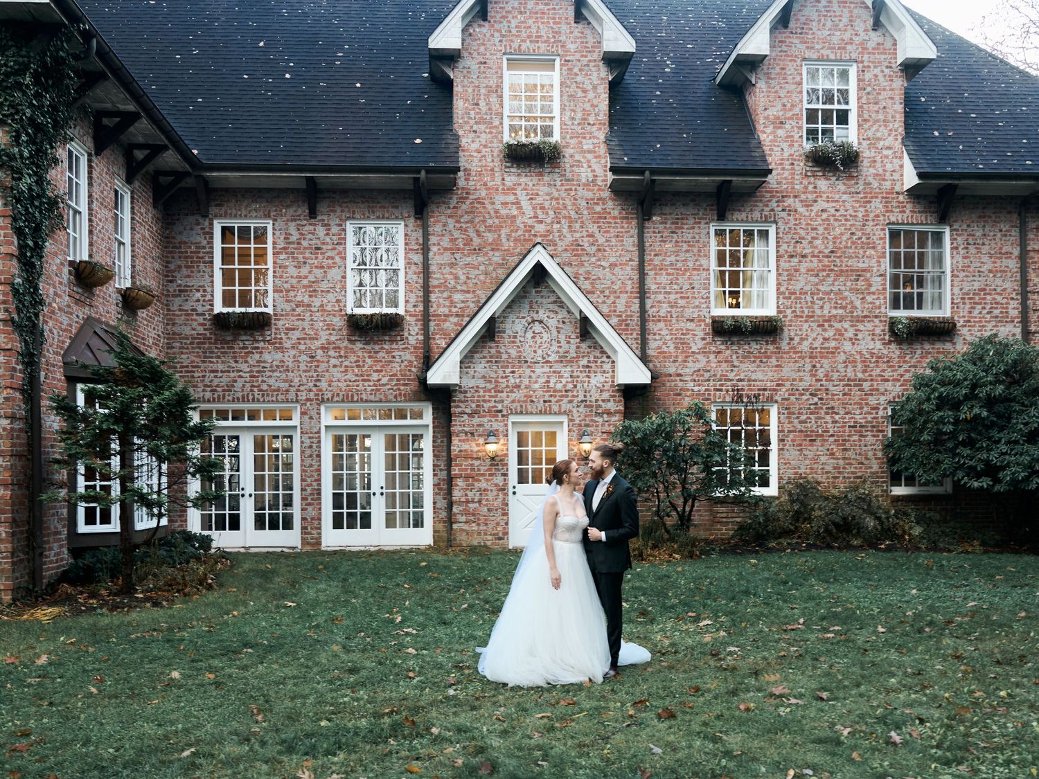 Couple posing for wedding photos in front of snow-dusted brick estate.
