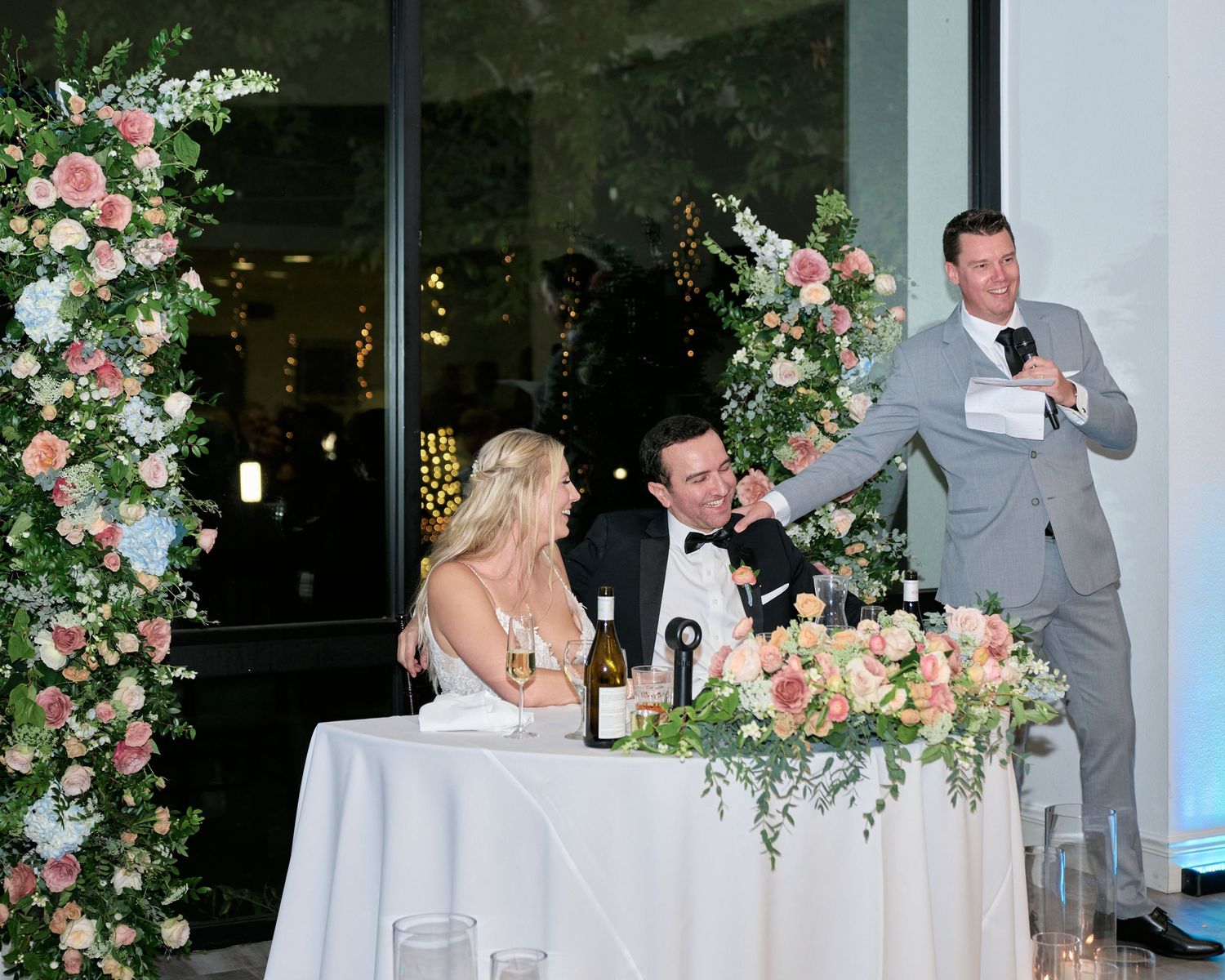 A wedding reception setup with floral arches and a sweetheart table during toasts.