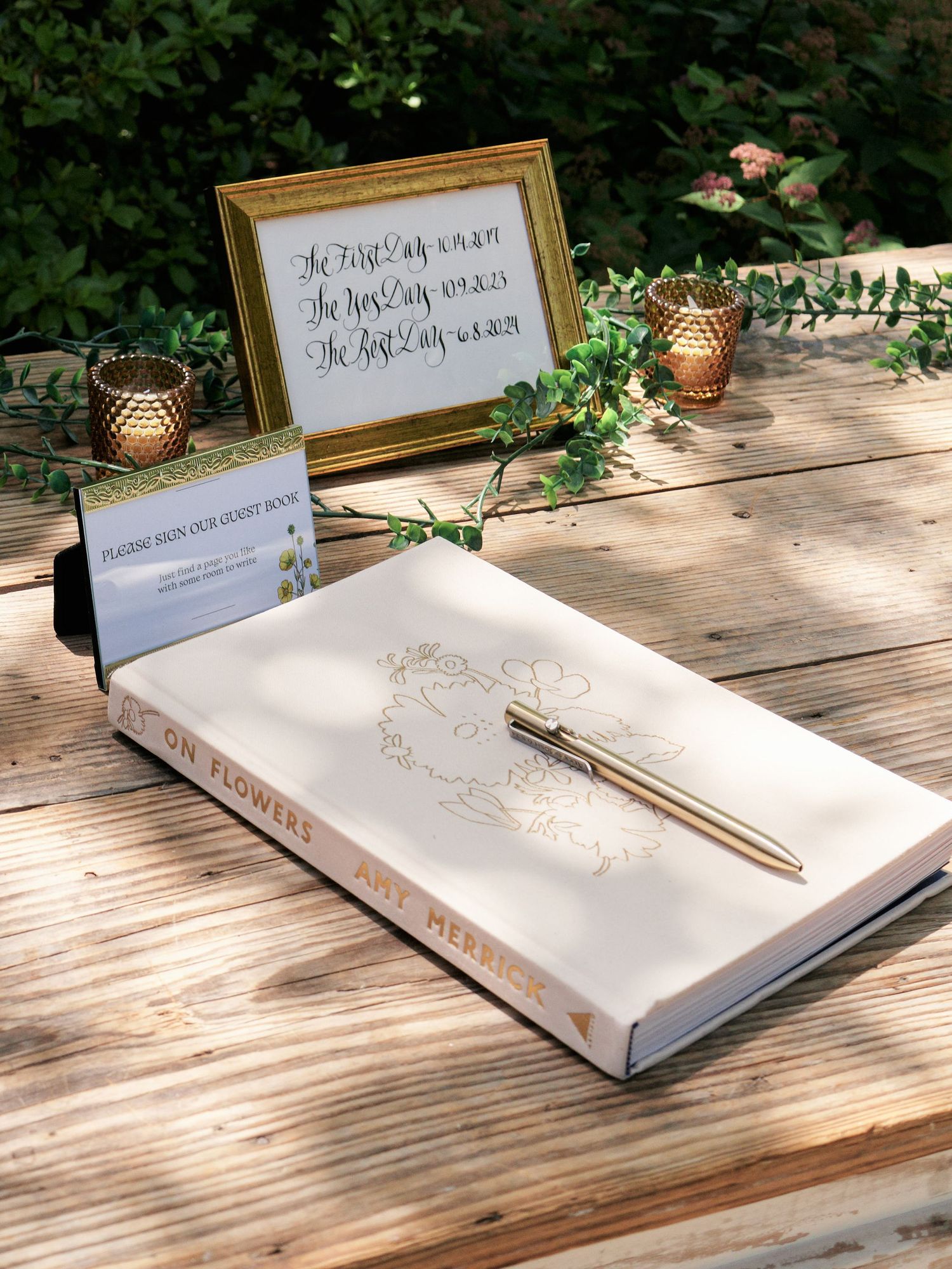 A wedding guest book and decorative frame sit on a wooden table with gold candle holders and greenery.