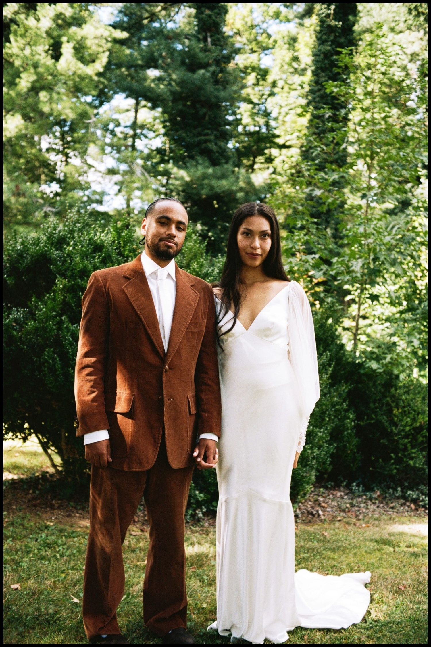 Newlyweds pose for formal portrait outdoors with bride in white gown and groom in brown suit.