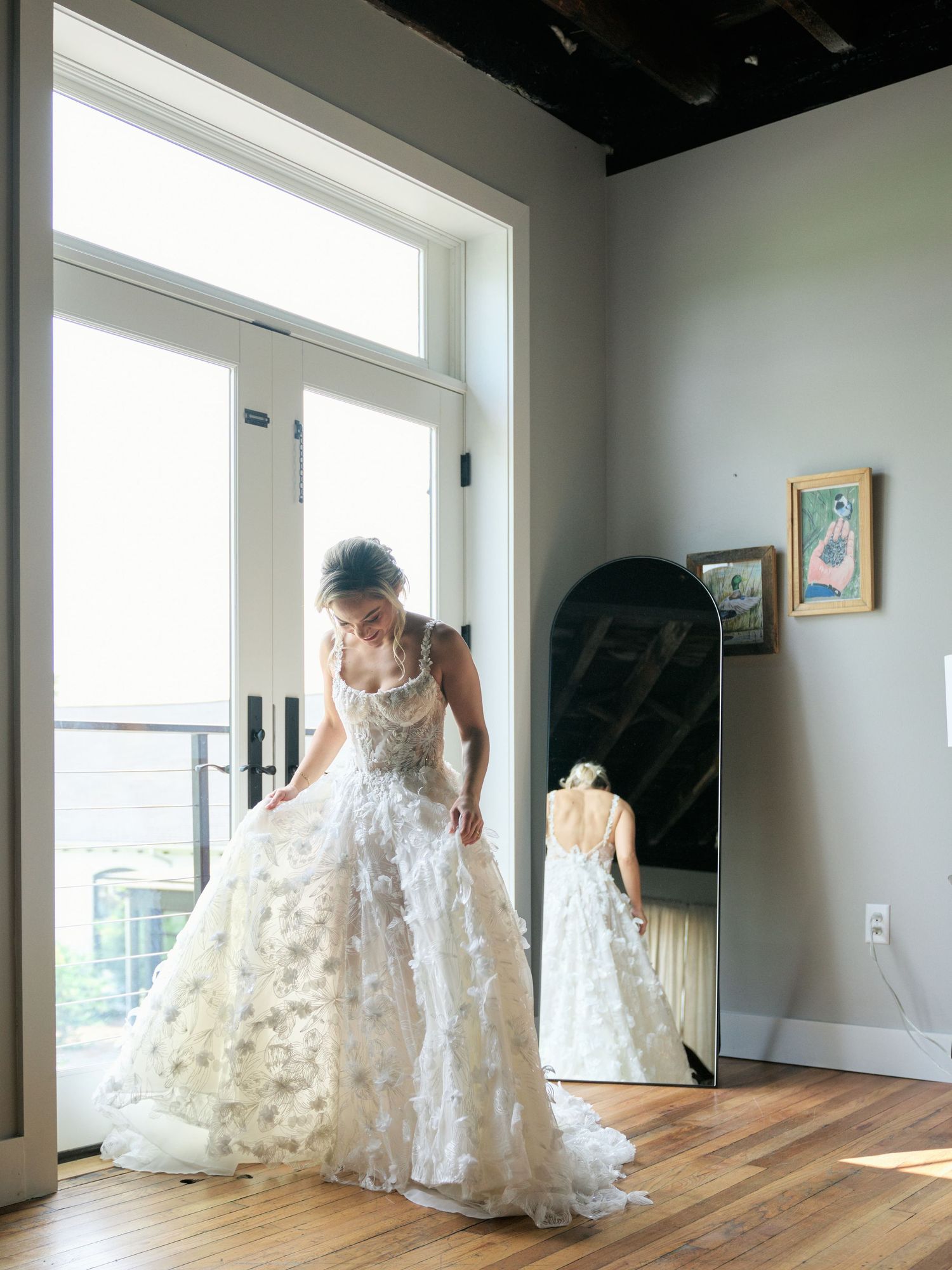 A bride in a flowing white floral lace wedding gown stands by sunlit windows with her reflection visible in a full-length mirror.