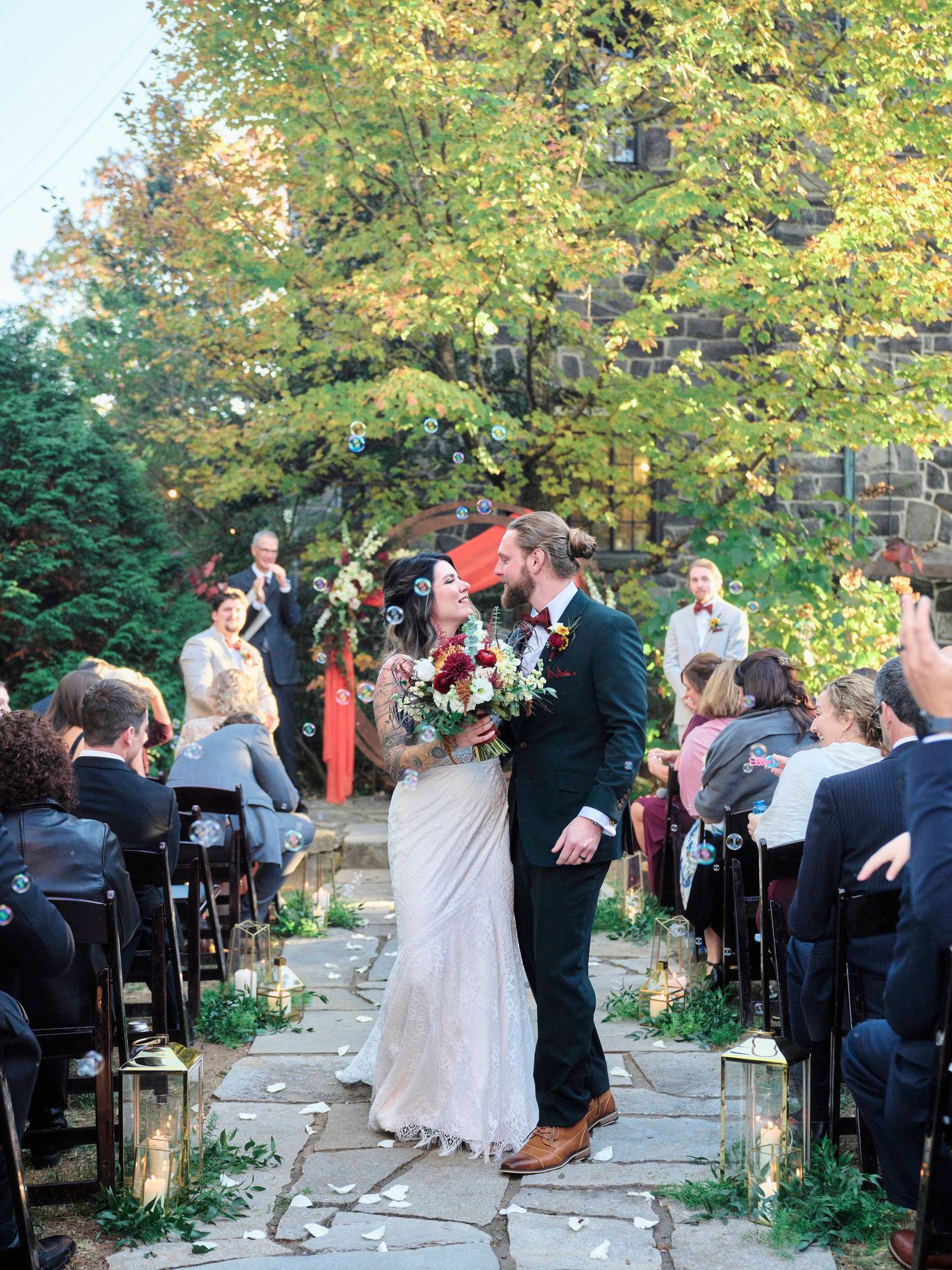 A wedding ceremony takes place on a stone path surrounded by fall foliage and seated guests.