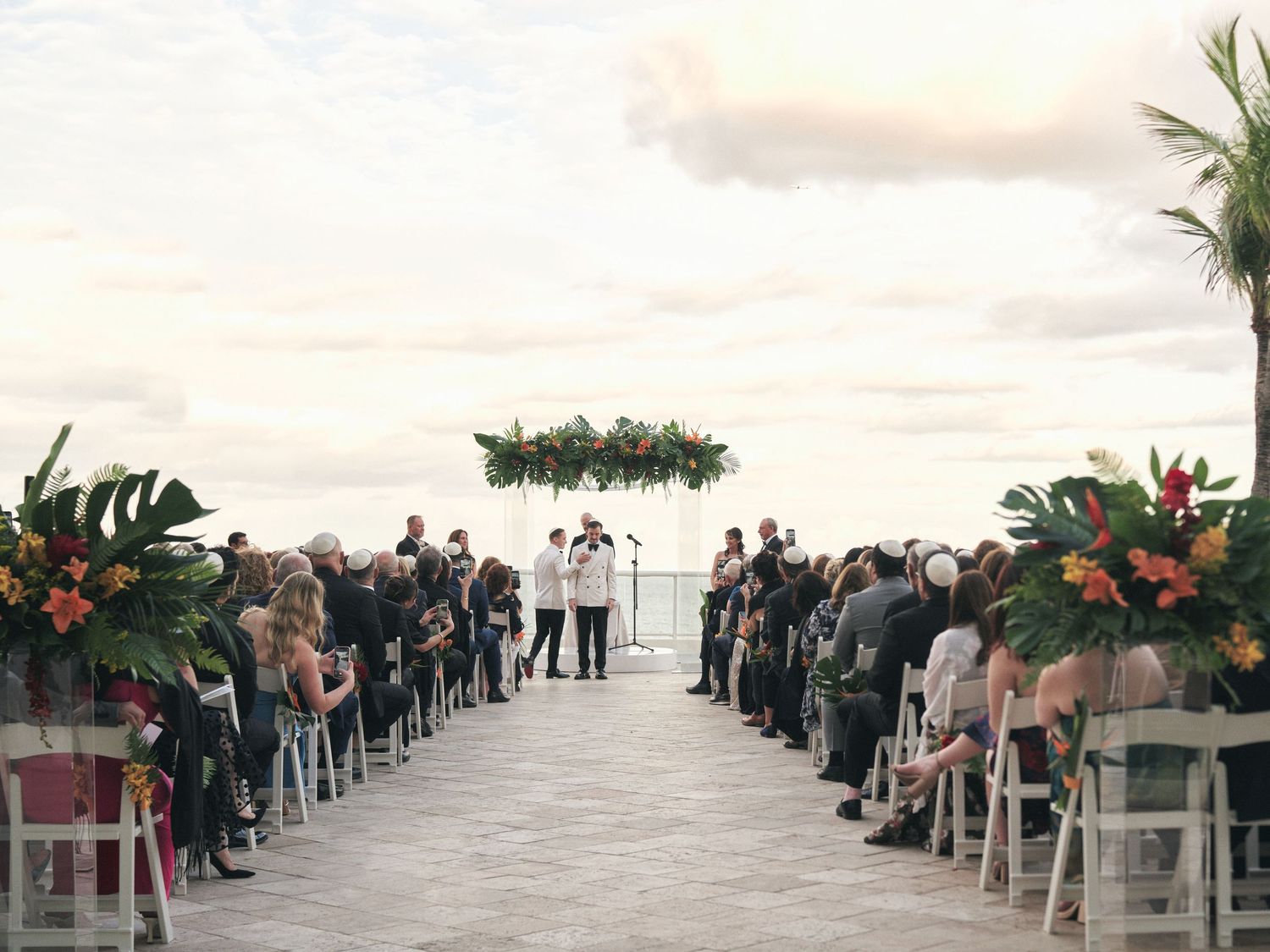 Outdoor wedding ceremony setup with floral arch and rows of white chairs on oceanfront deck.