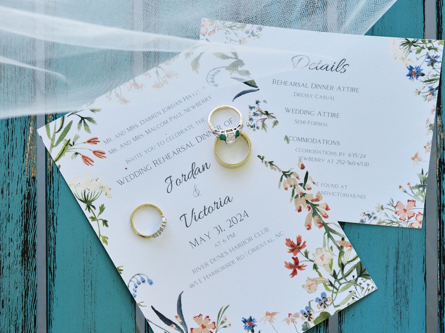 Wedding invitation with floral design and gold wedding rings displayed on turquoise wooden surface.