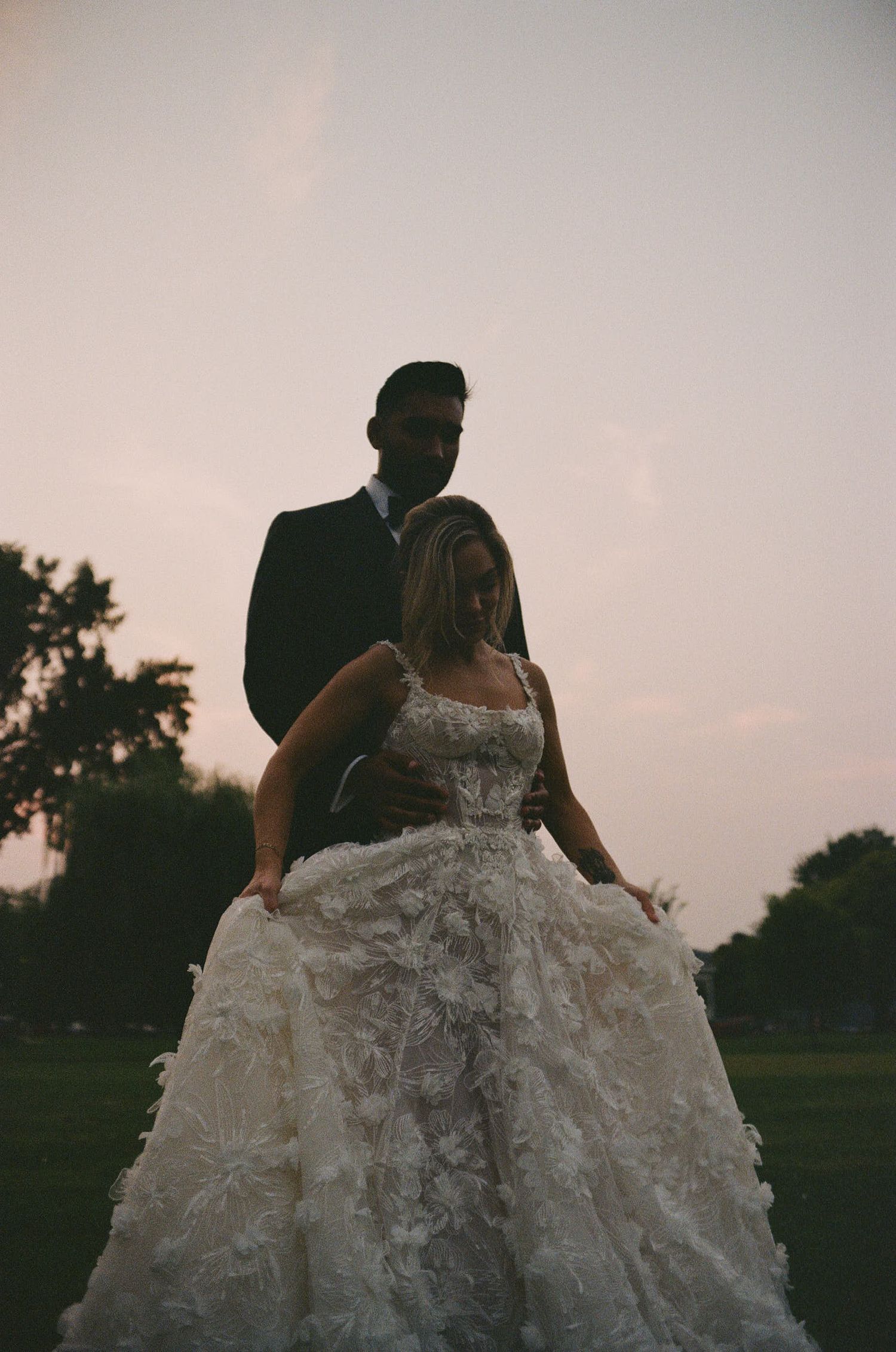 A silhouetted couple stands in a grassy field at dusk, with the bride wearing a detailed lace wedding gown.