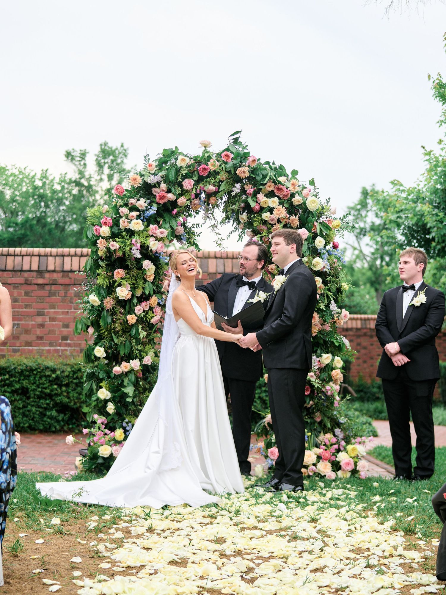 Couple exchanging vows under a lush floral arch with pink and cream roses during outdoor ceremony.