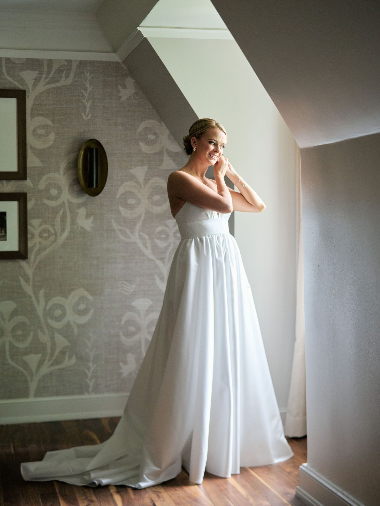 A person in a white wedding gown stands in natural light by a window, putting on jewelry.