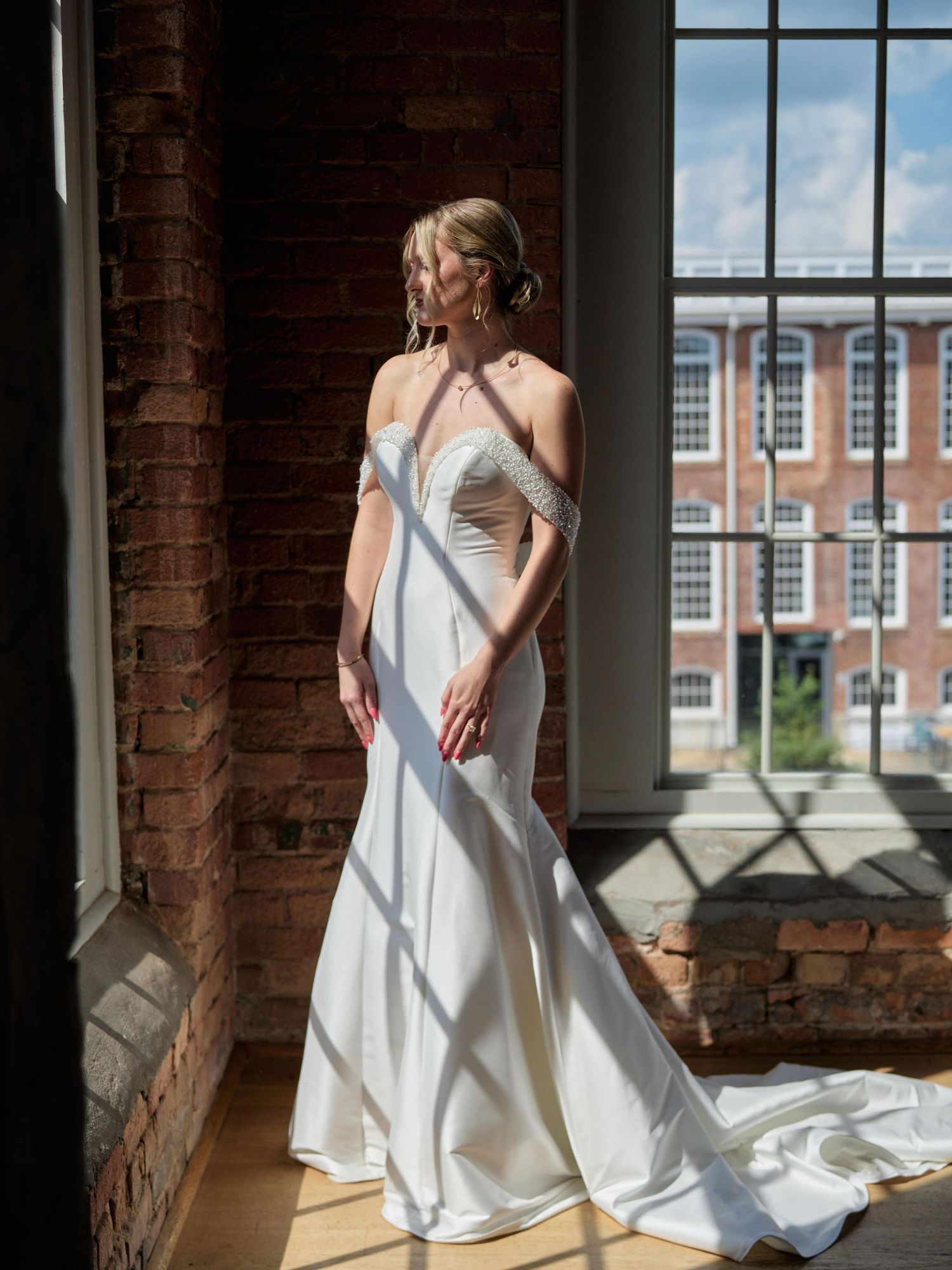 Elegant bride in fitted white gown poses by window with exposed brick and city view.
