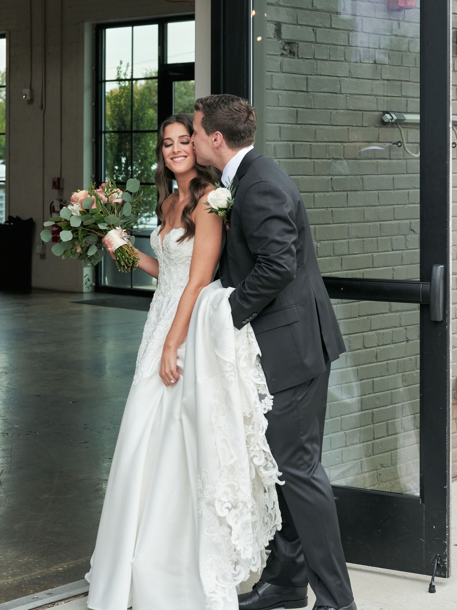 A romantic wedding moment with a couple in formal attire standing by a brick wall, featuring a white lace gown and bouquet.