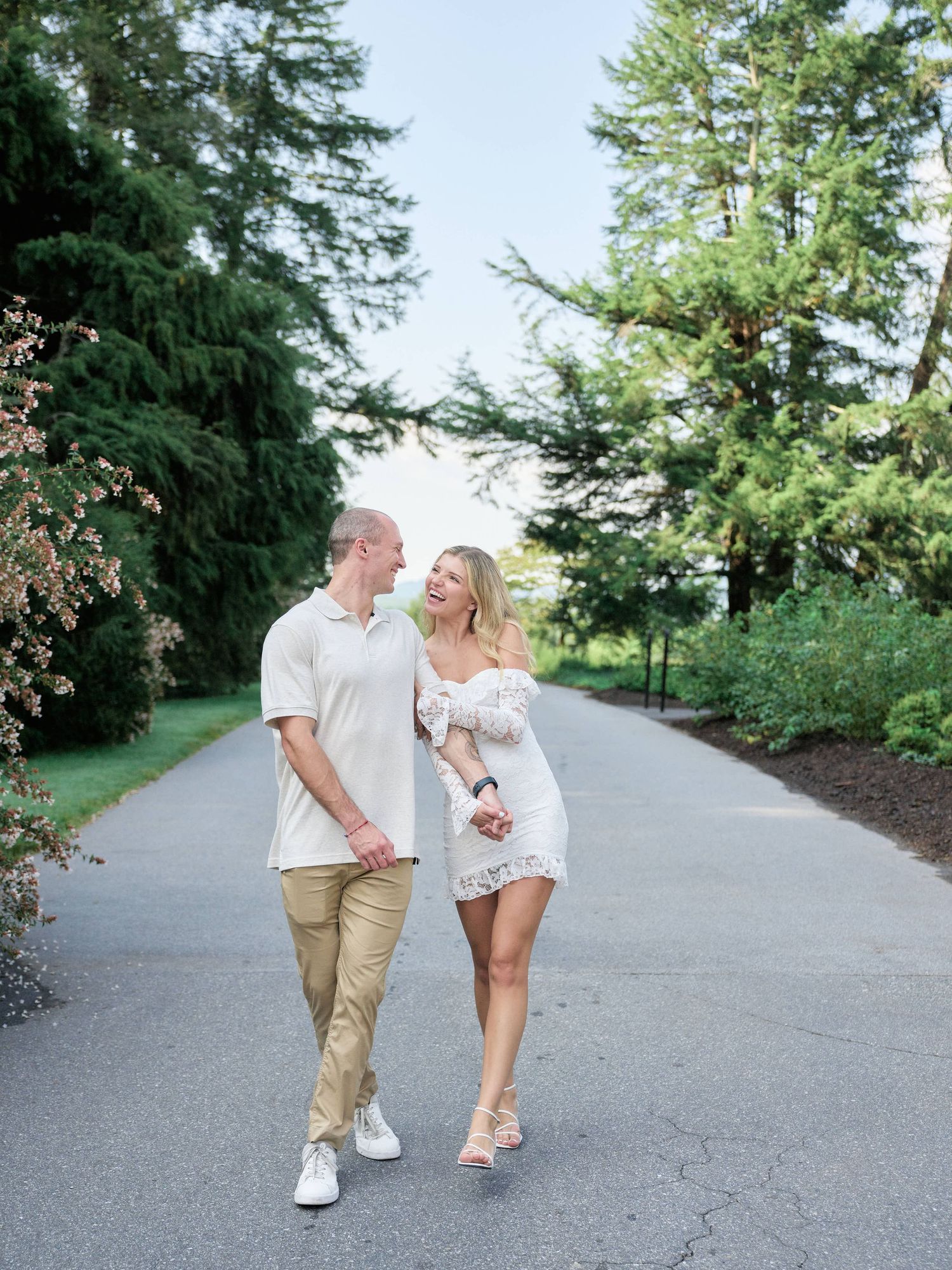 A couple walking and laughing together on a paved path surrounded by lush green trees on a sunny day.