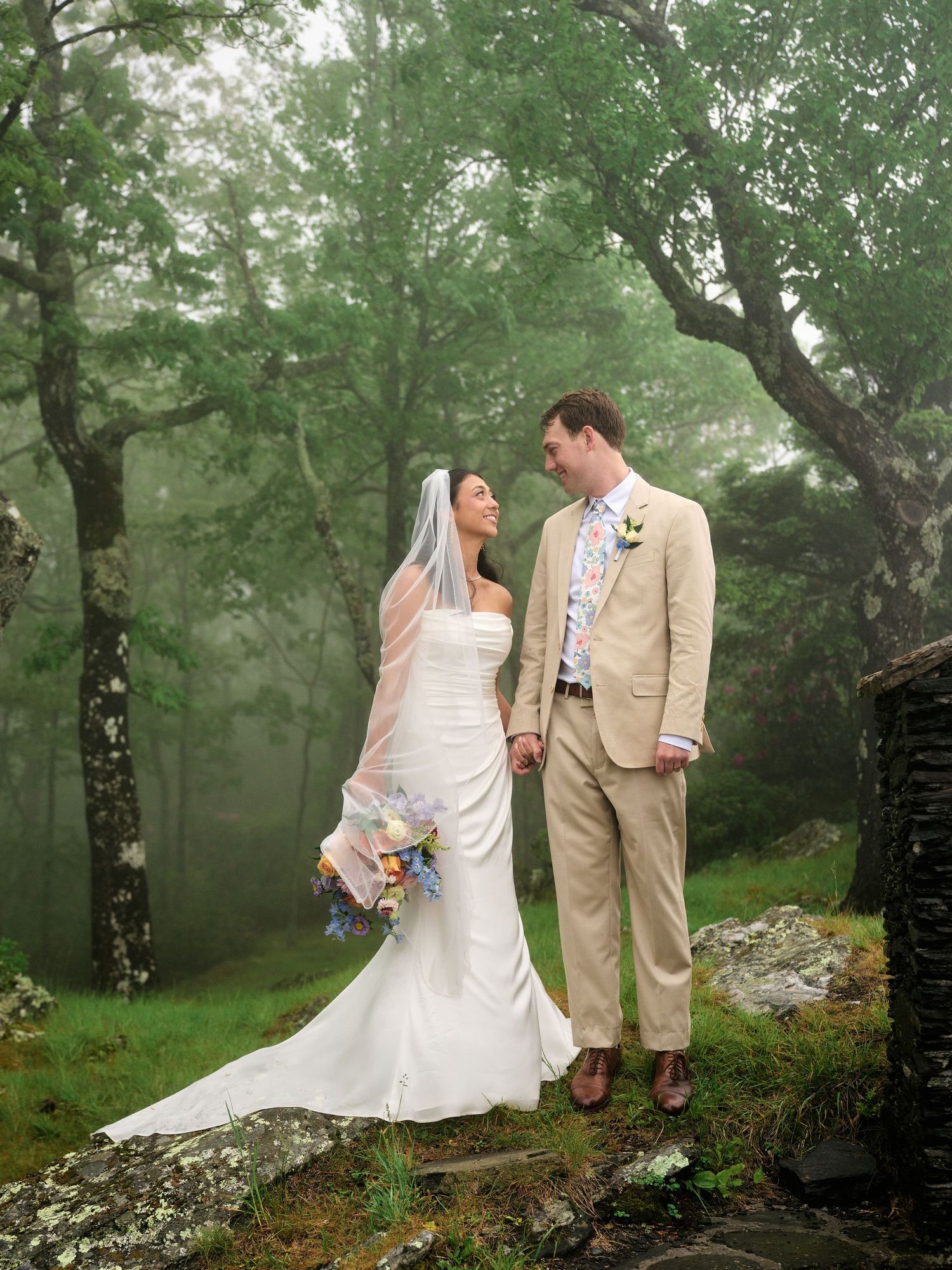 Bride and groom hold hands in misty forest, she in white gown with veil, he in tan suit with floral tie.