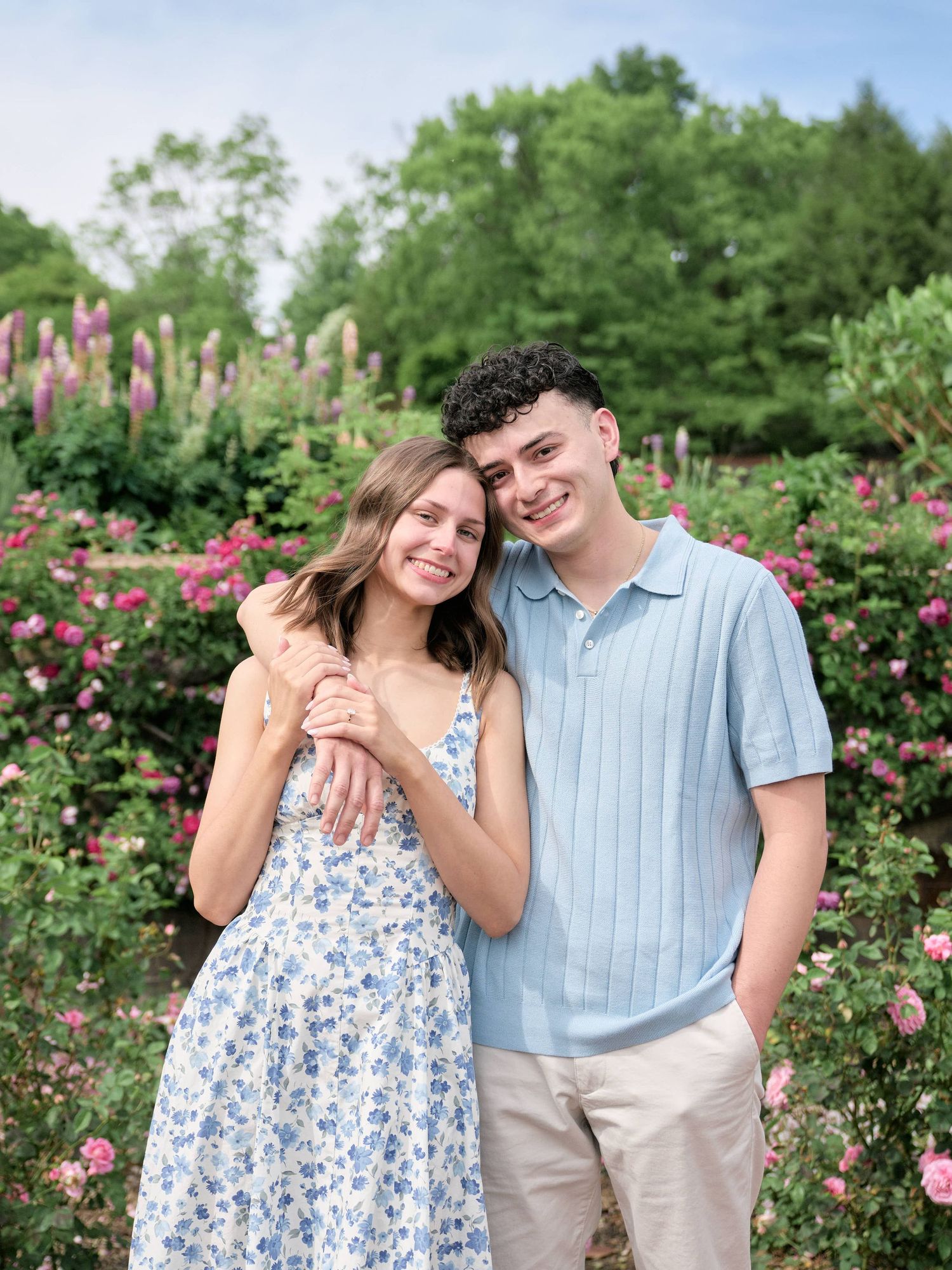 A smiling couple poses together in a blooming garden, the woman in a floral dress and man in a blue polo shirt.
