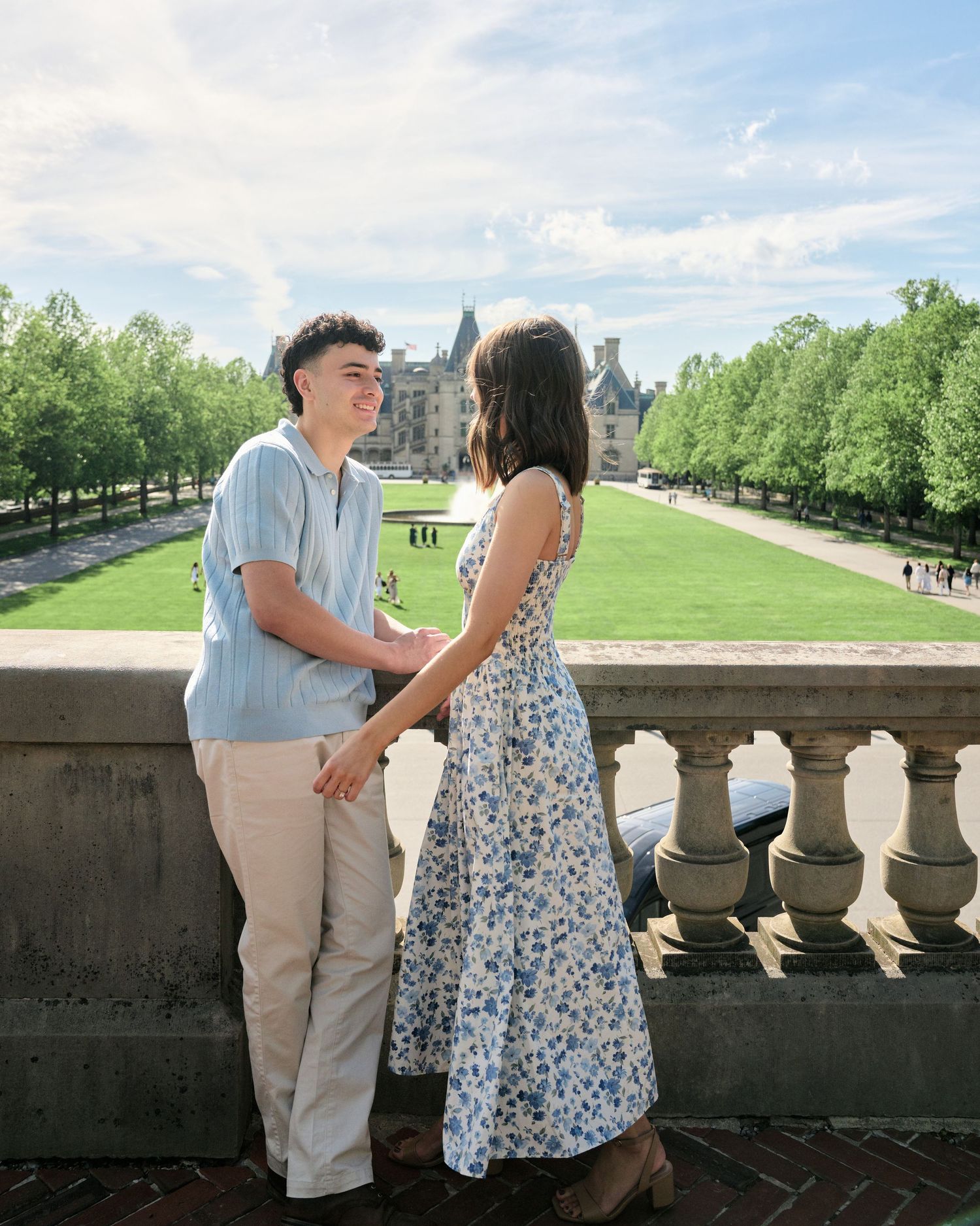Couple holding hands on a stone balcony at Biltmore Estate, surrounded by lush green trees and manicured gardens.