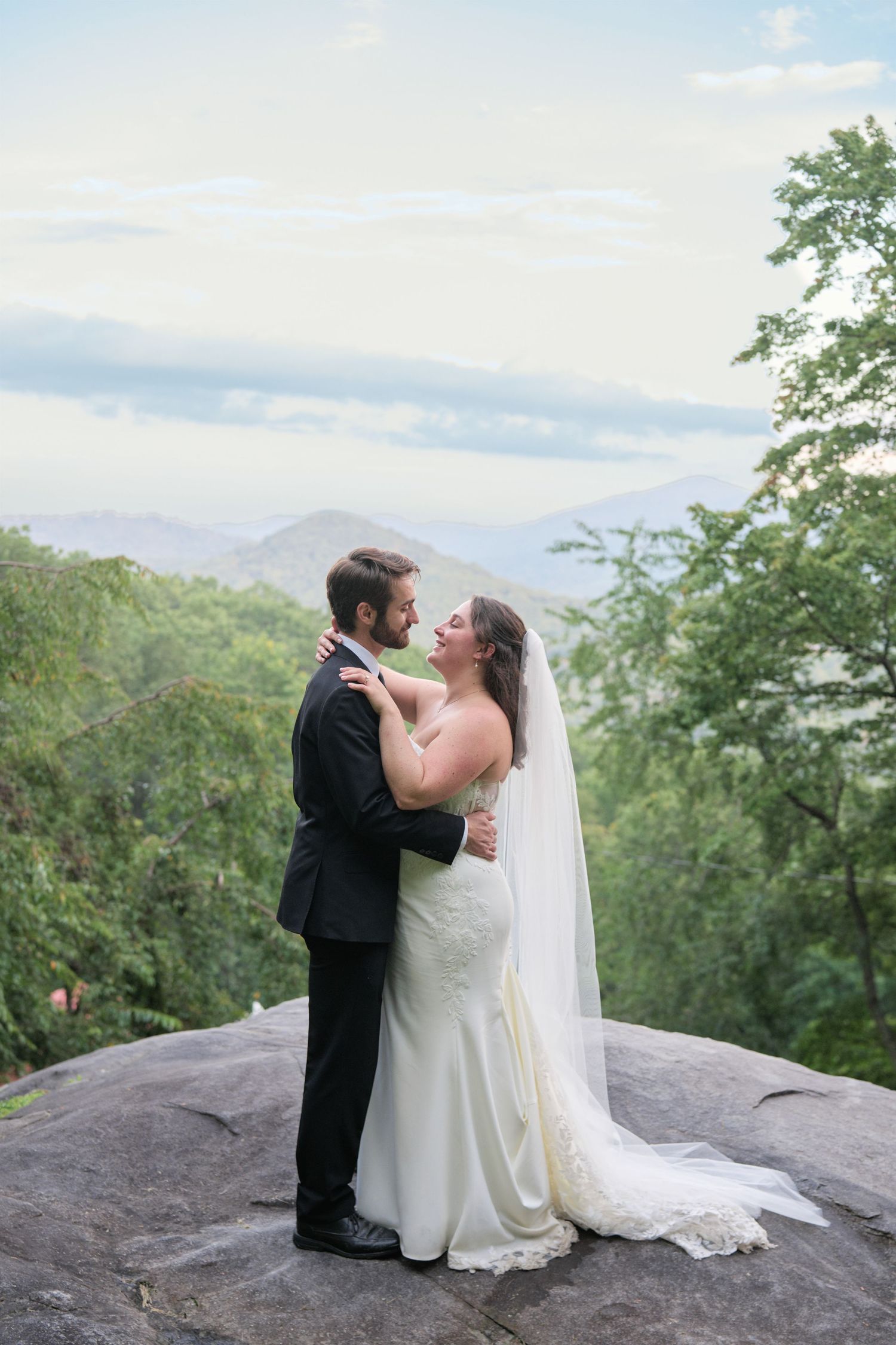 Bride and groom embrace on a rocky overlook with lush green mountains and cloudy sky backdrop on their wedding day.