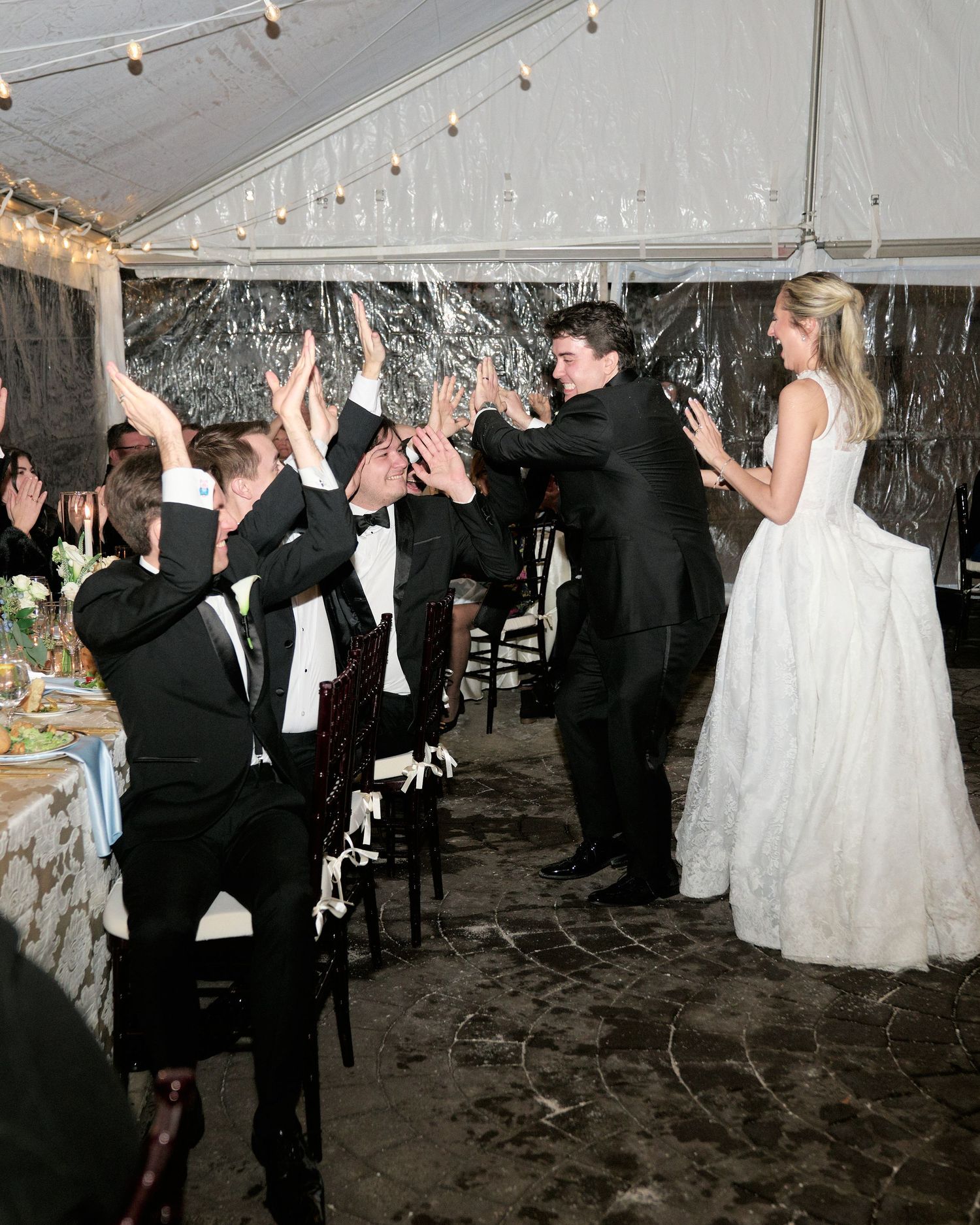Bride in white gown high-fives groomsmen in black tuxedos under a decorated tent at a wedding reception.