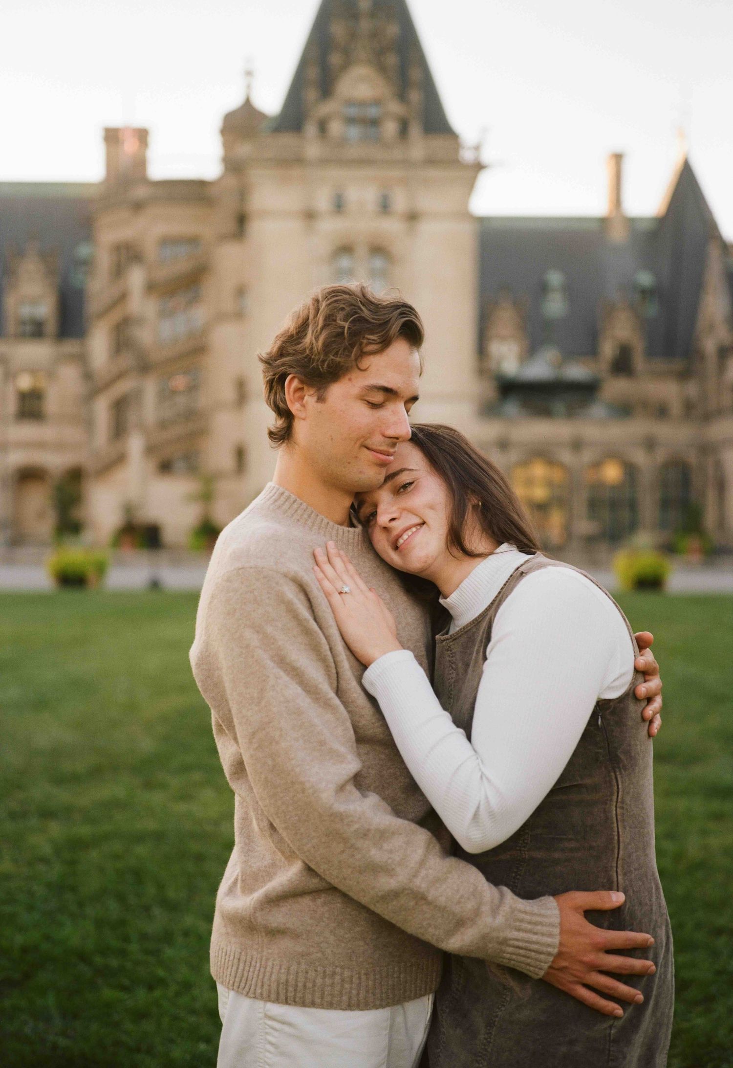 Couple embracing romantically in front of Biltmore Estate at golden hour, woman smiling with eyes closed.