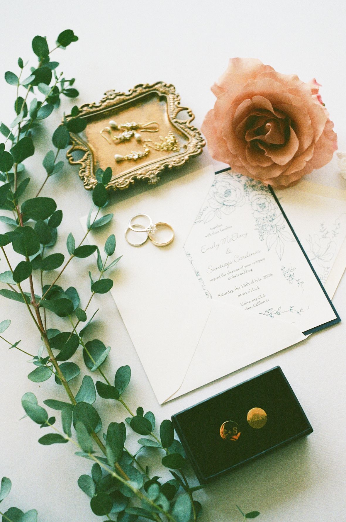 A romantic arrangement featuring a golden frame, rings, and a peach-colored rose on a wooden surface with greenery.