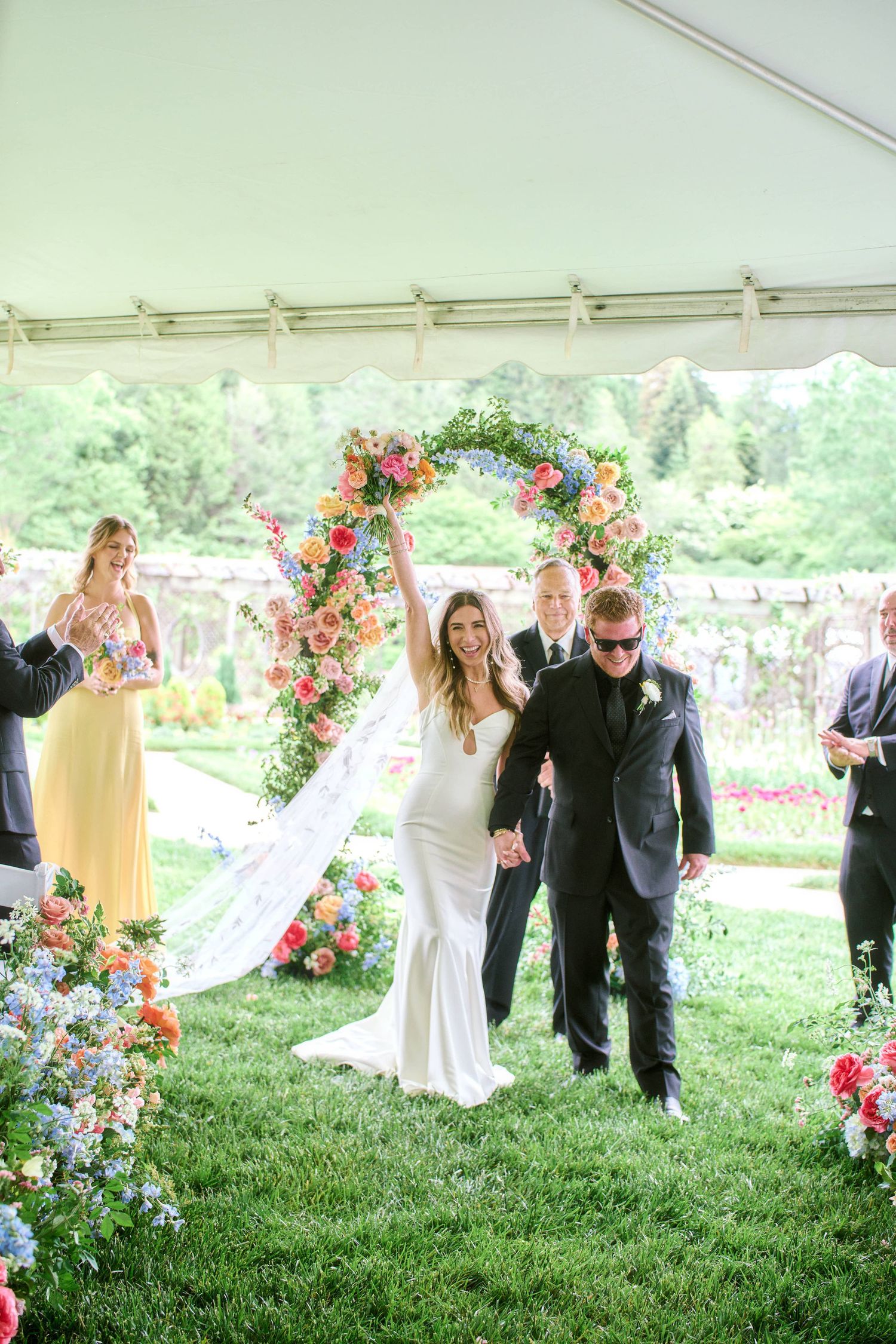 A wedding ceremony setup outdoors with a floral arch and guests gathered on a grassy lawn under a white tent.