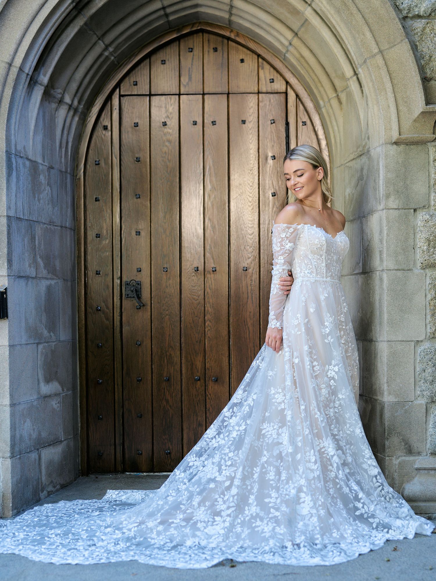 Stunning off-shoulder lace wedding dress with cathedral train posed by arched wooden door.