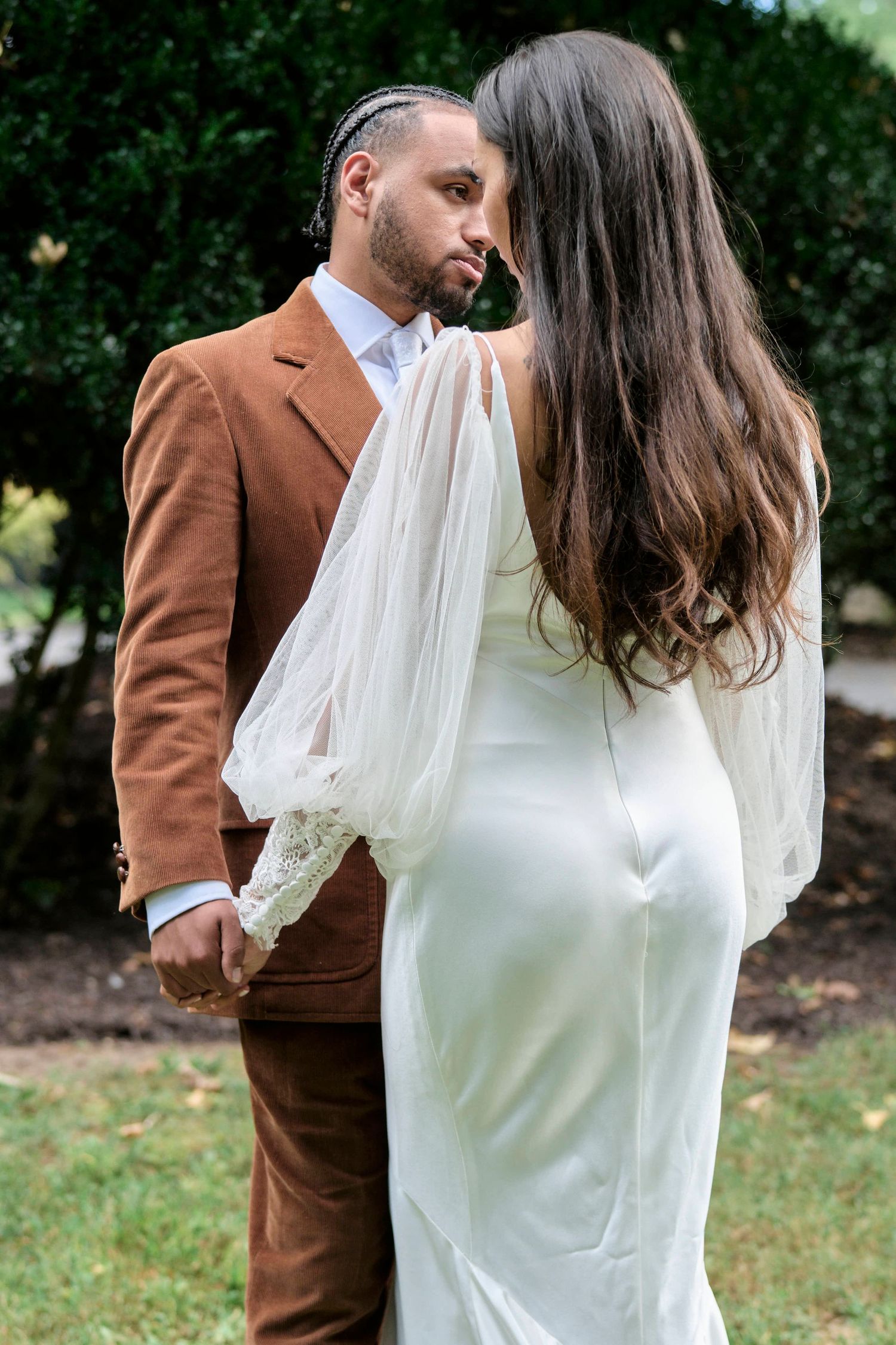 A couple in brown suit and white dress share an intimate moment in a garden setting.
