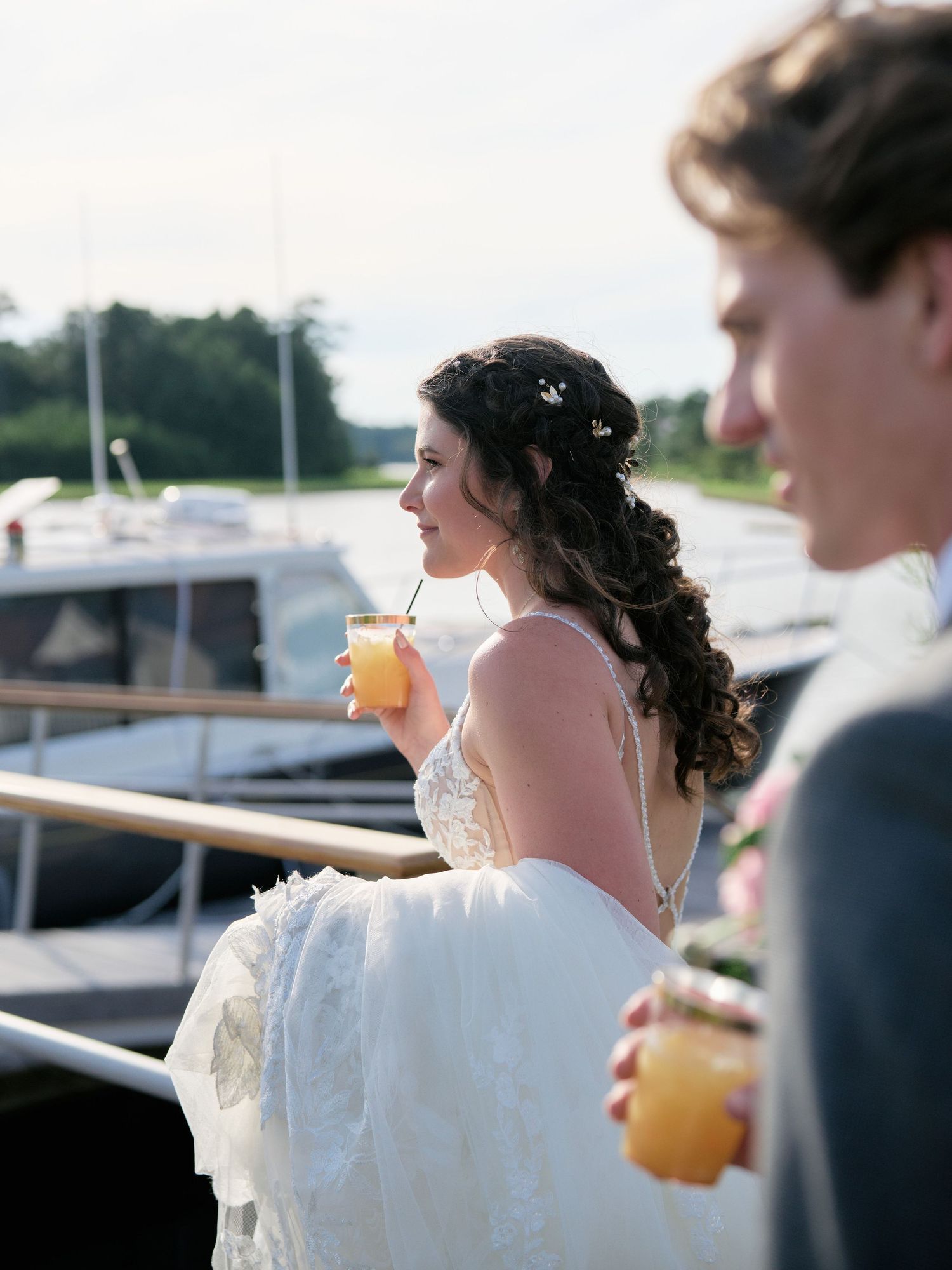 Couple in wedding attire enjoying orange drinks together on a boat dock during a summer celebration.