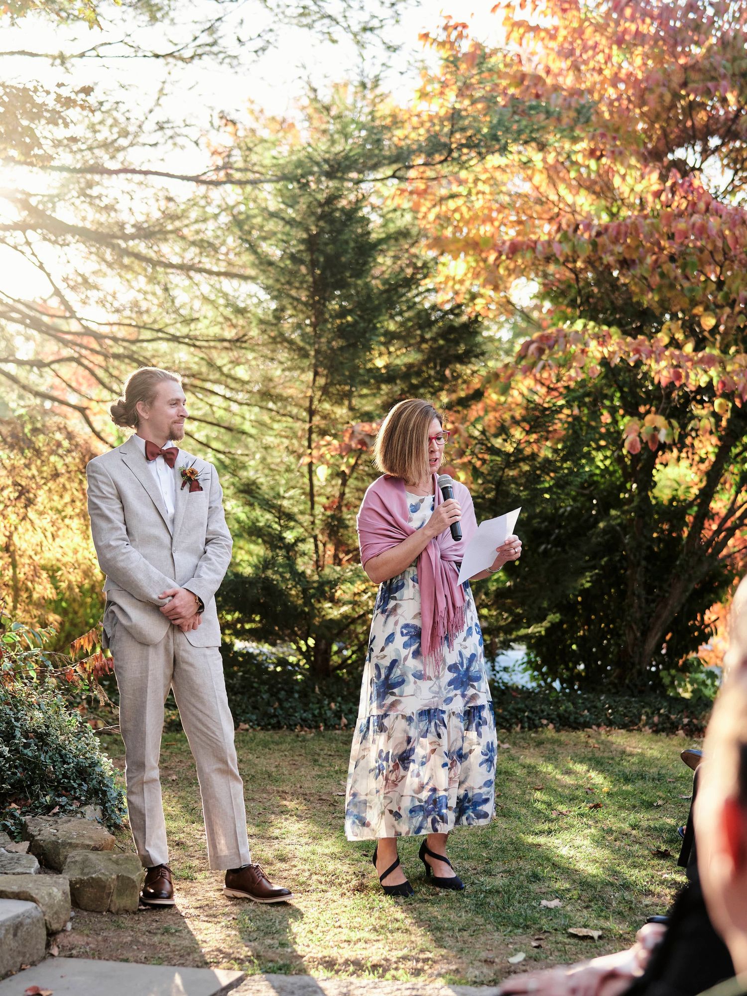 Wedding speakers stand on stone steps against a backdrop of autumn trees with golden sunlight.