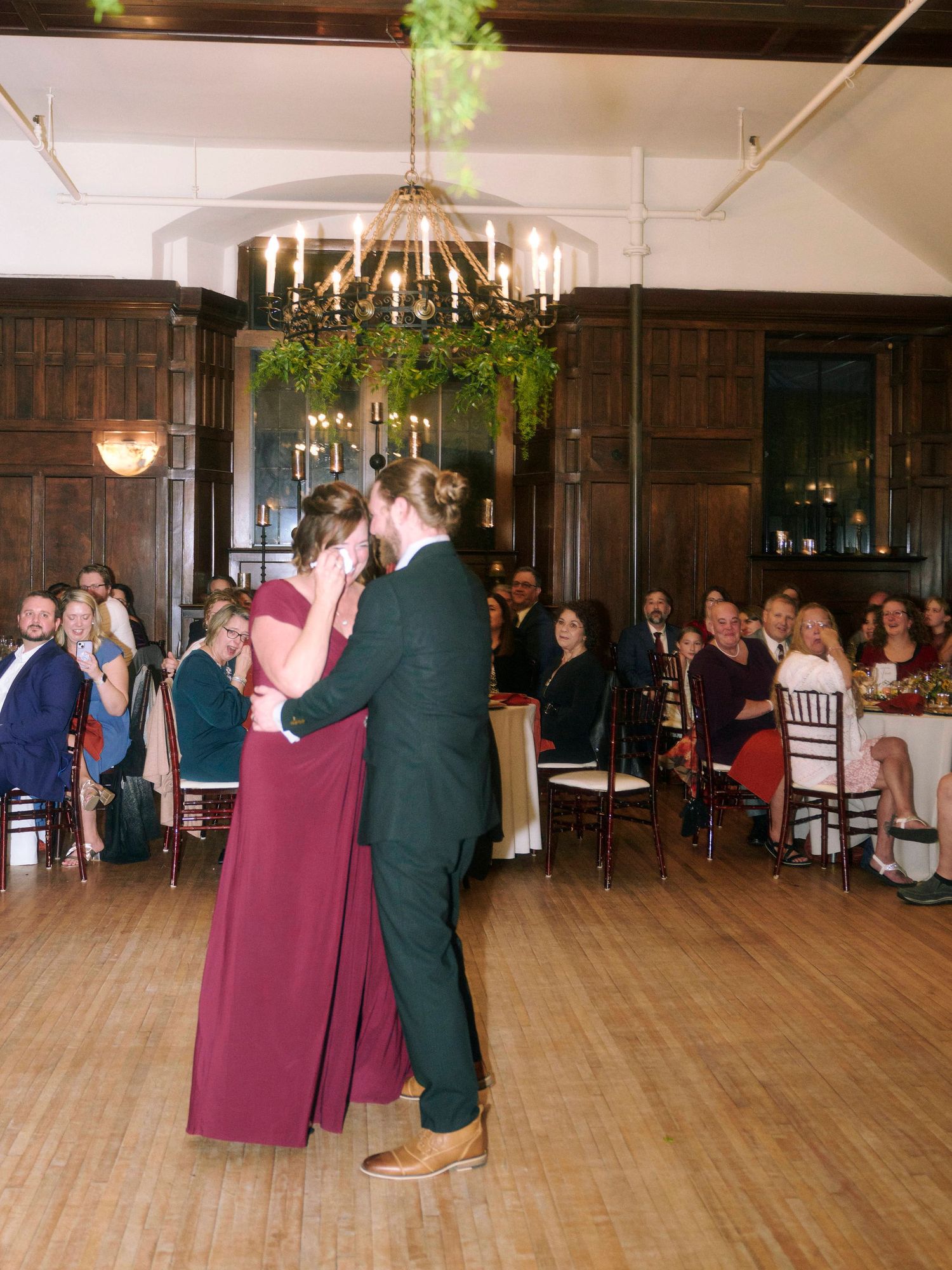 Couples dancing at wedding reception in elegant wood-paneled venue with chandelier and greenery.