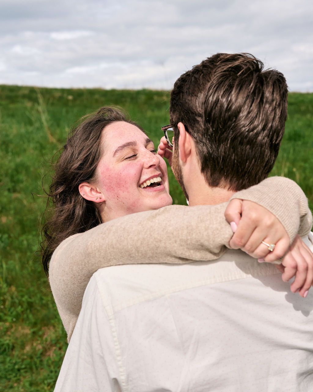 A joyful couple in beige sweaters embraces and laughs together in a green field under cloudy skies.
