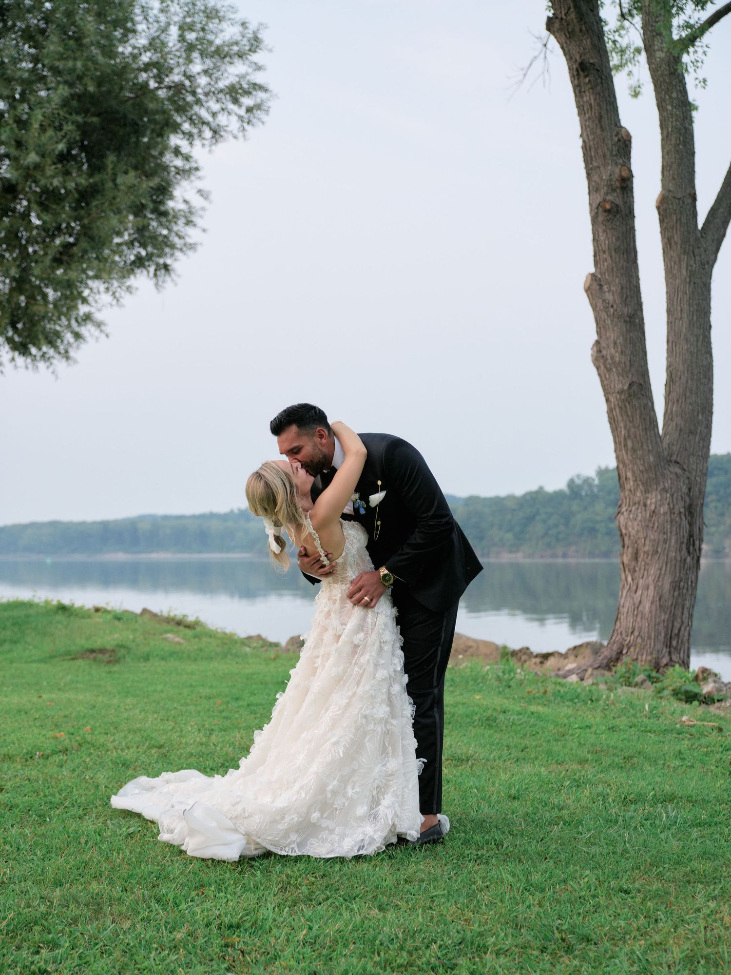 A romantic dip-pose wedding photo captured on a green lawn overlooking a serene lake with tall trees in the background.