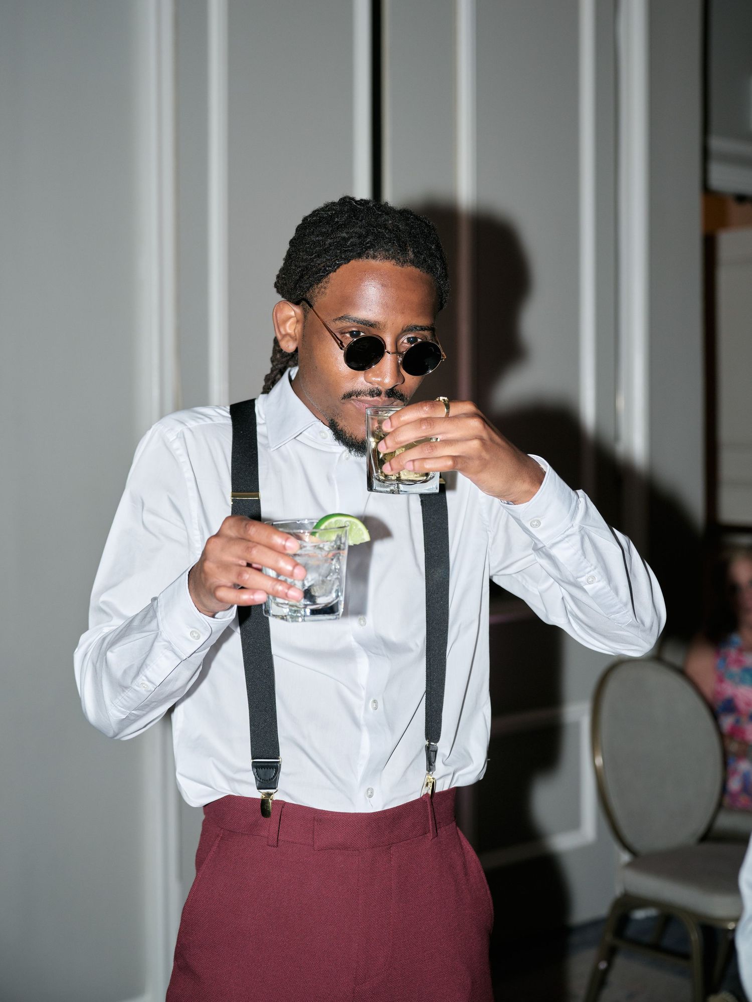A stylish person in suspenders and sunglasses enjoying two cocktails at a formal event.