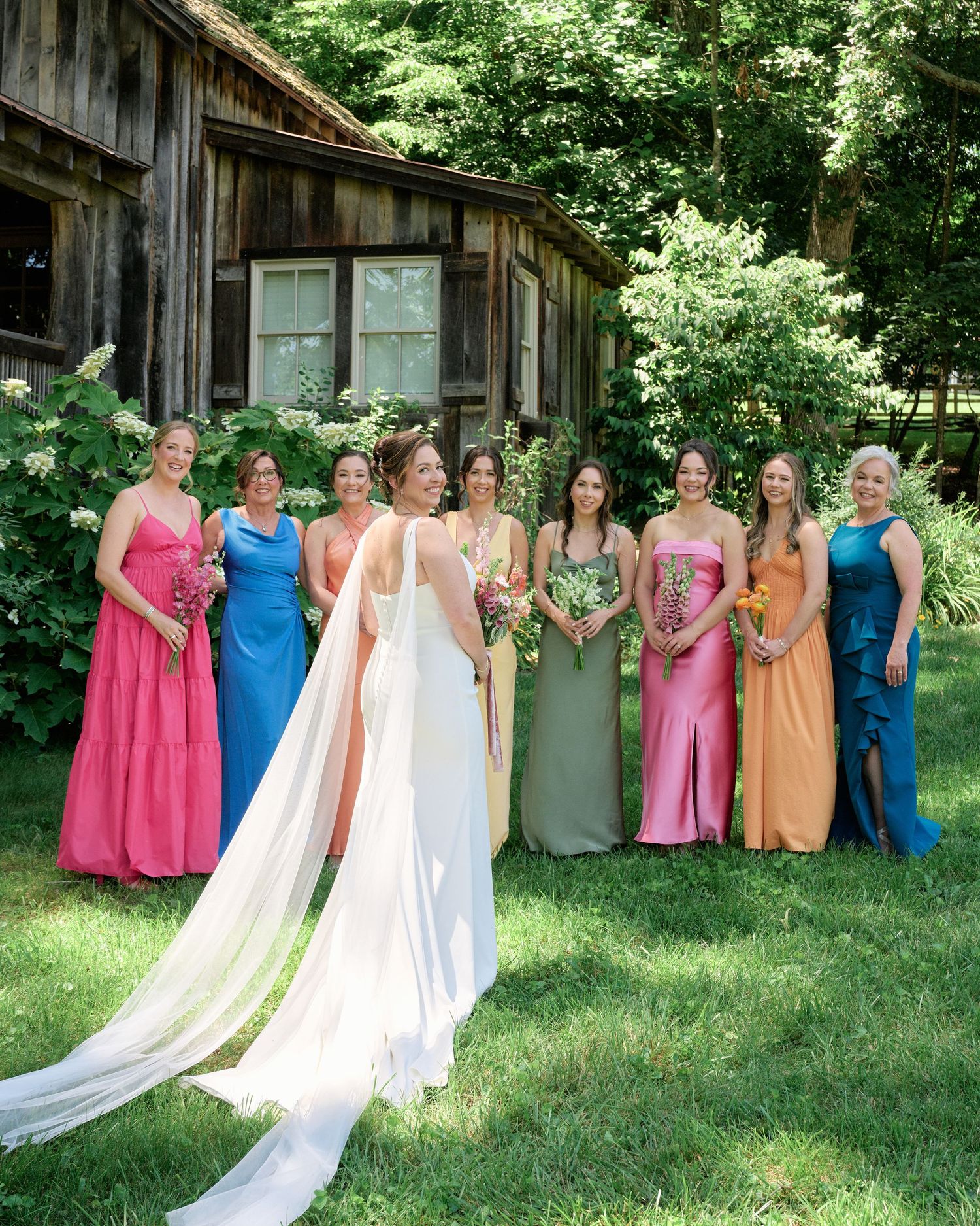 A bridal party in rainbow-colored dresses stands with the bride in front of a wooden cabin.