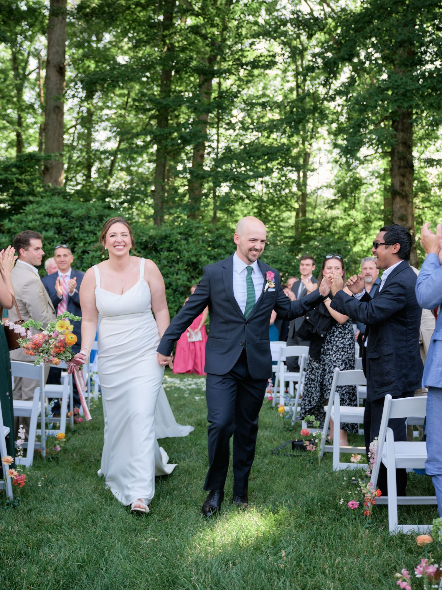 Newlyweds walk back down the aisle while wedding guests cheer and clap in an outdoor woodland setting.