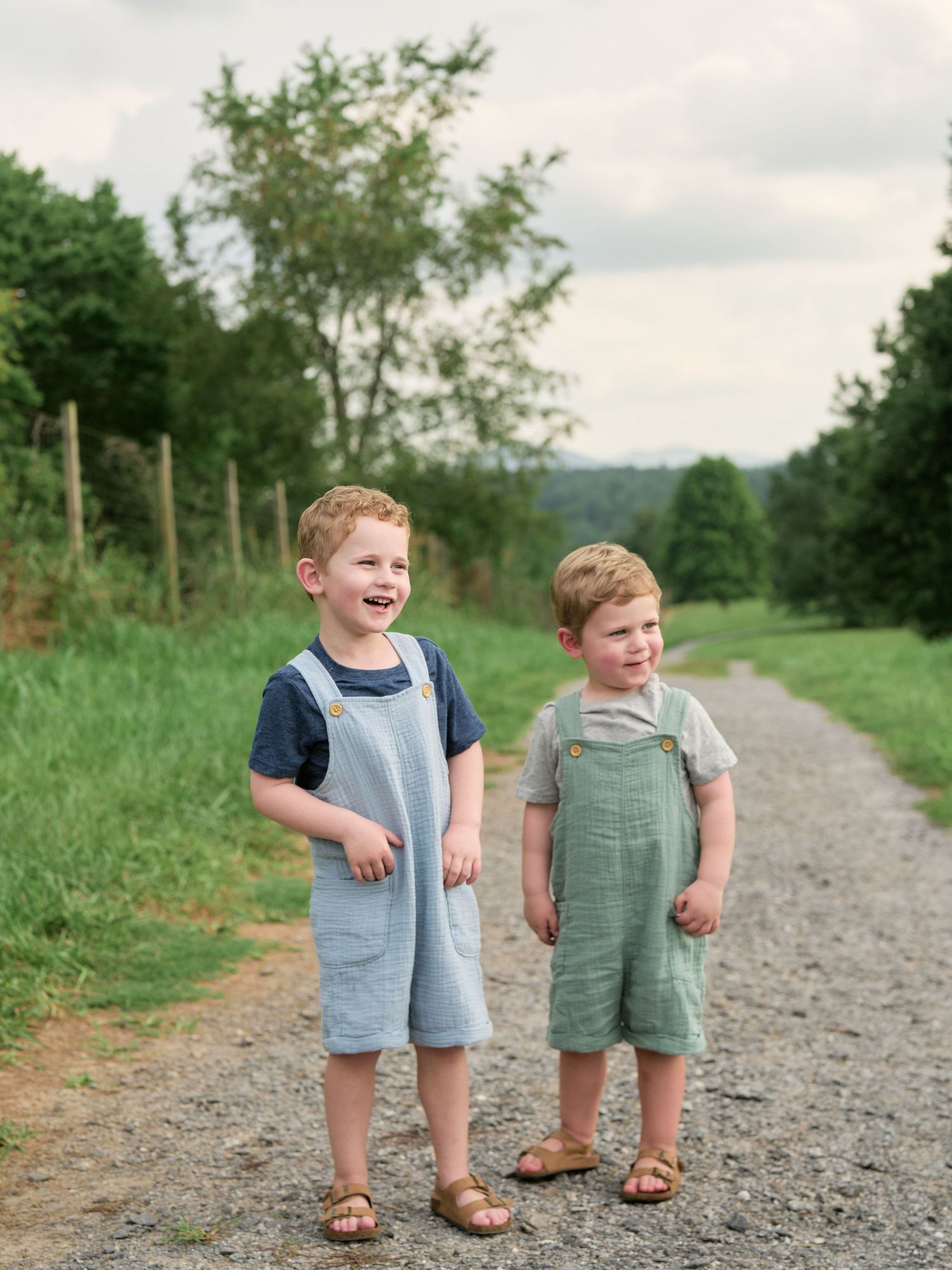Two children in casual summer outfits walking together on a gravel path surrounded by lush greenery.