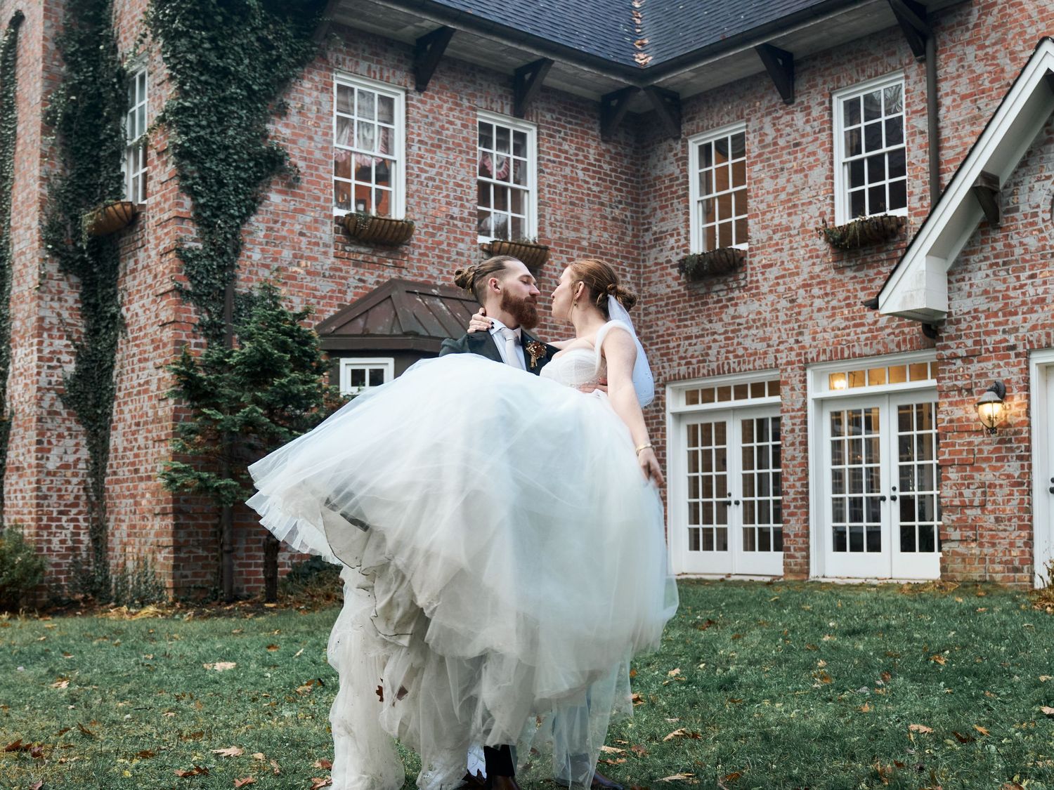 Joyful wedding couple embracing in front of historic brick mansion with ivy-covered walls.