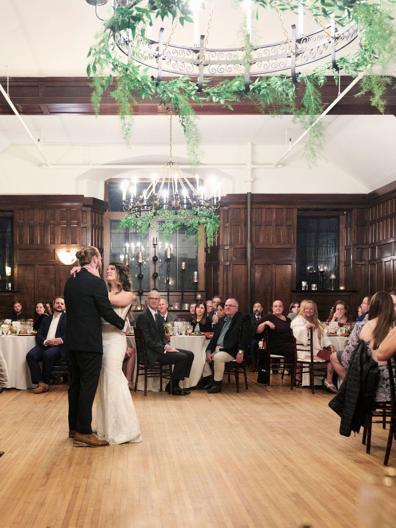 A couple shares their first dance in an elegant wood-paneled reception hall with hanging greenery and chandelier.