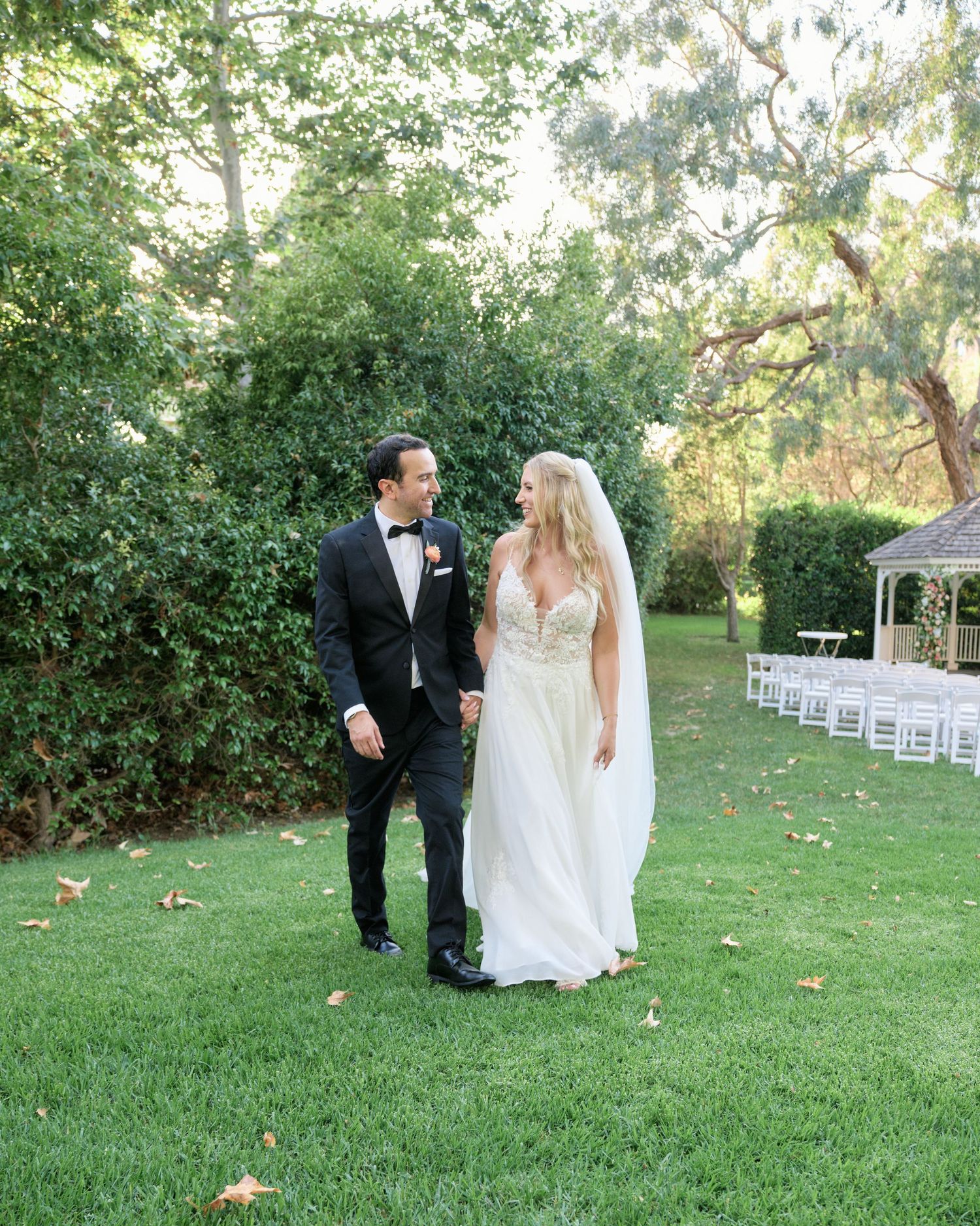 A bride and groom share a candid moment on a lush green lawn with white chairs set up for an outdoor wedding ceremony.