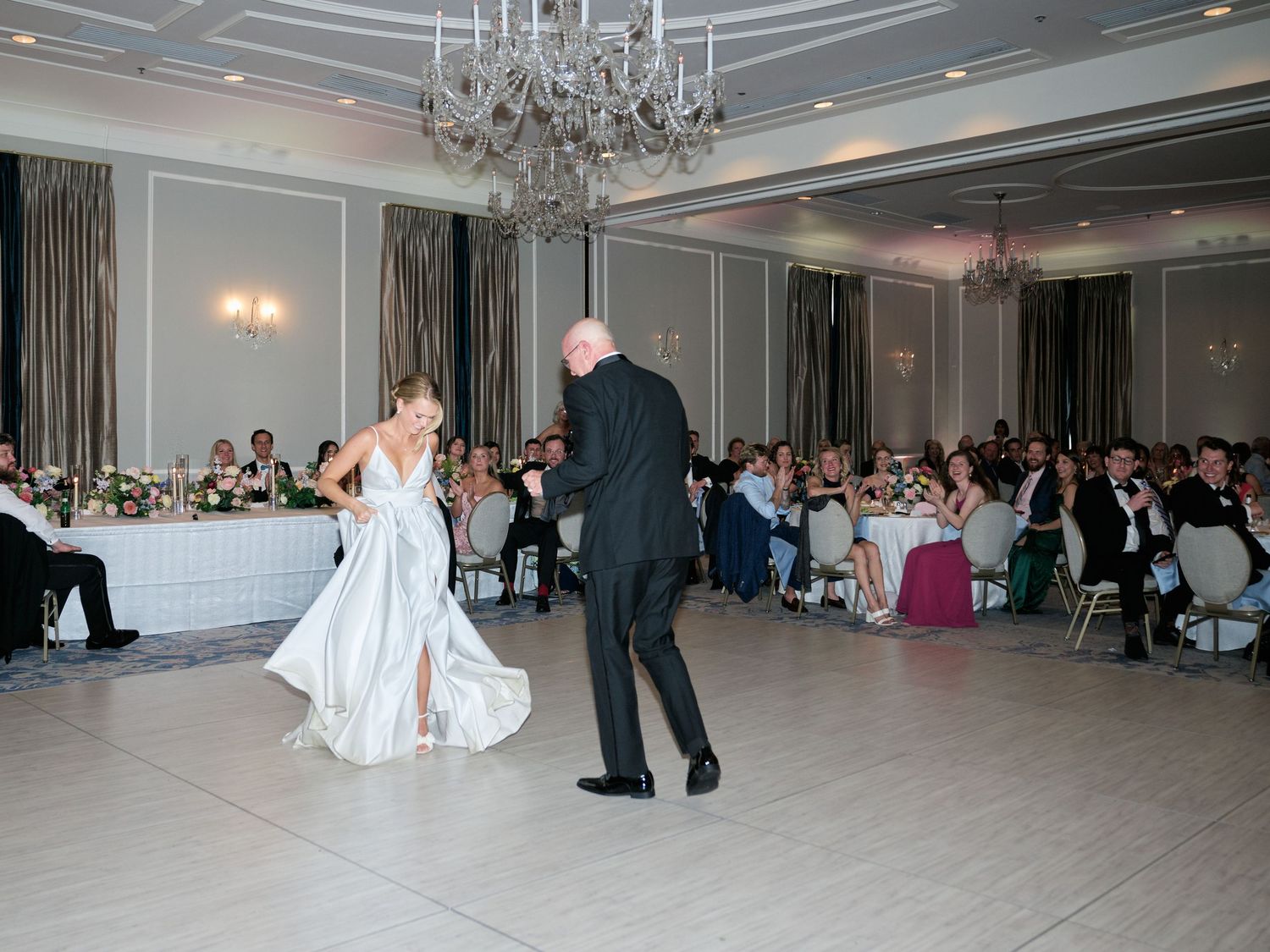 A bride and father dance in an elegant reception hall with guests seated at round tables.