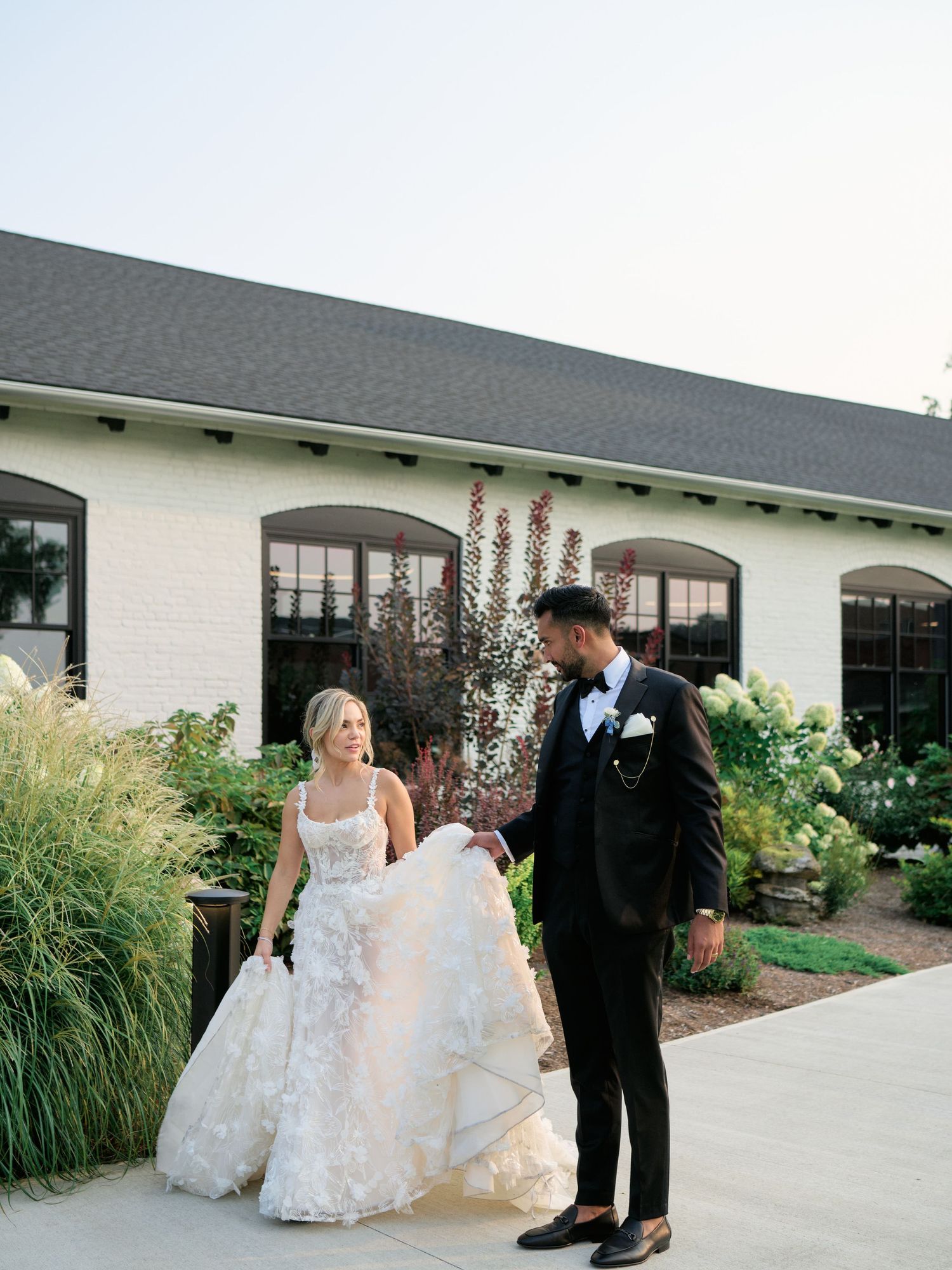 A bride in a flowing white dress and groom in a black tuxedo share a moment together on a garden pathway.