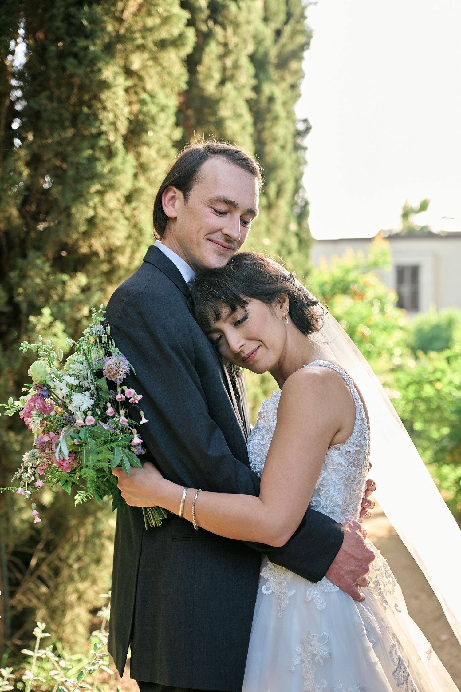 A tender wedding embrace captured in a sun-lit garden with cypress trees.