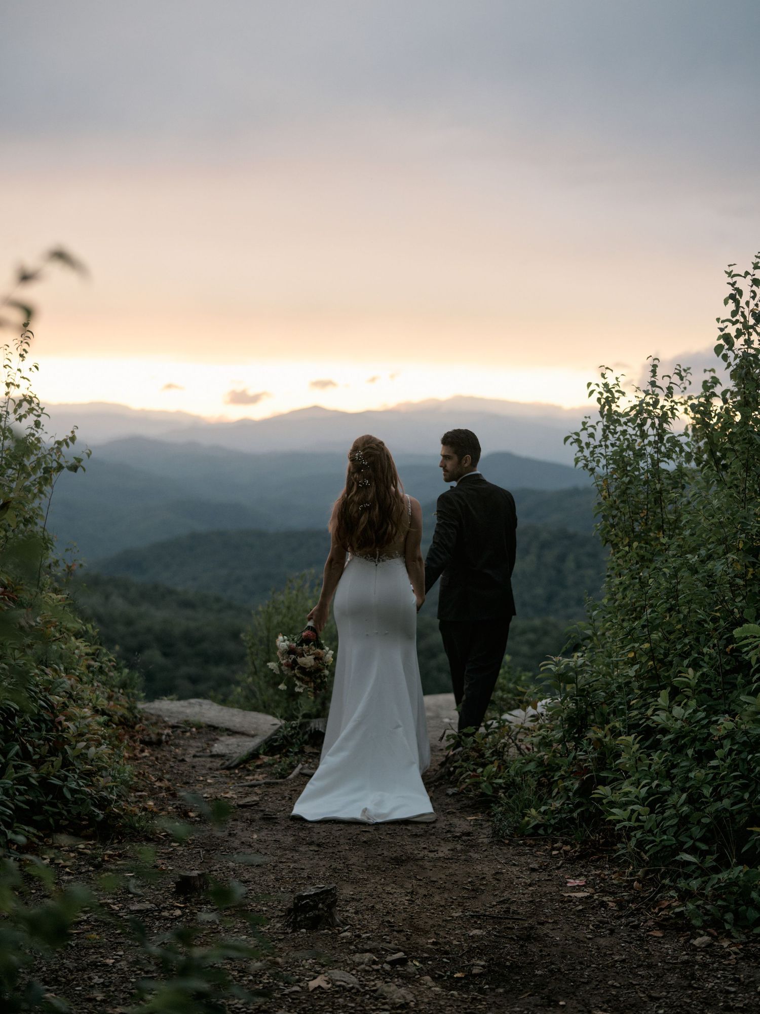 A couple in wedding attire stands on a mountain overlook during sunset, surrounded by lush greenery and mountain ranges.