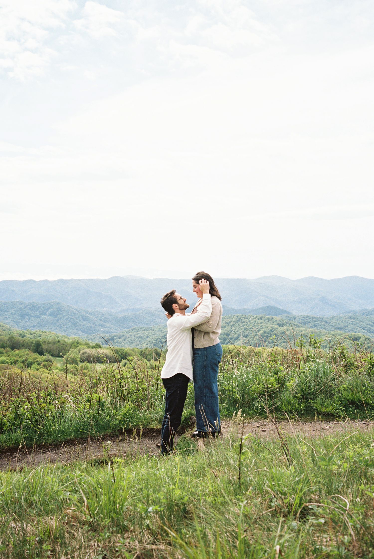 Couple embraces romantically in a scenic mountain meadow with rolling hills and lush greenery in the background.