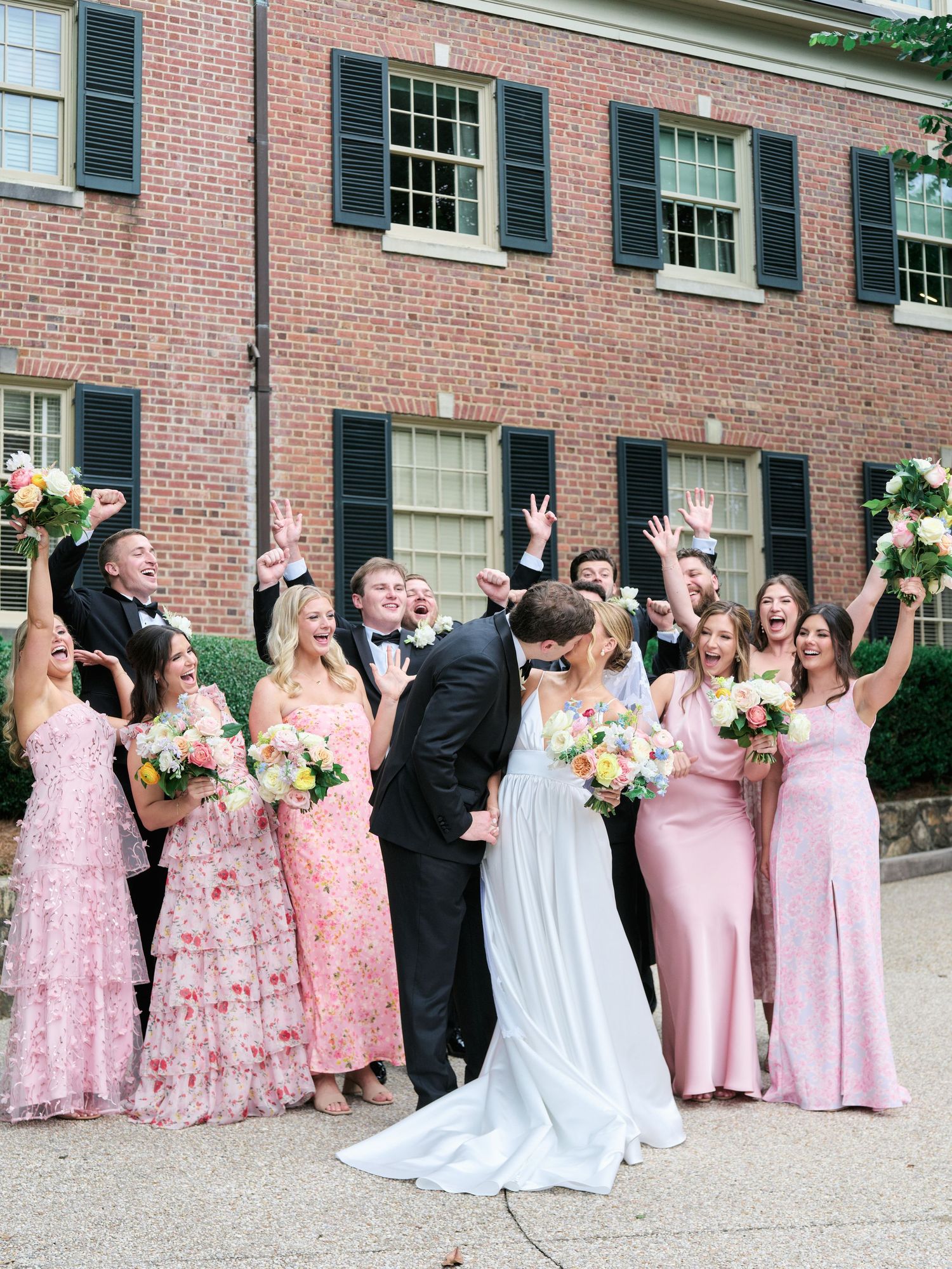 Wedding party celebrates with joyful poses outside brick venue with white trim.