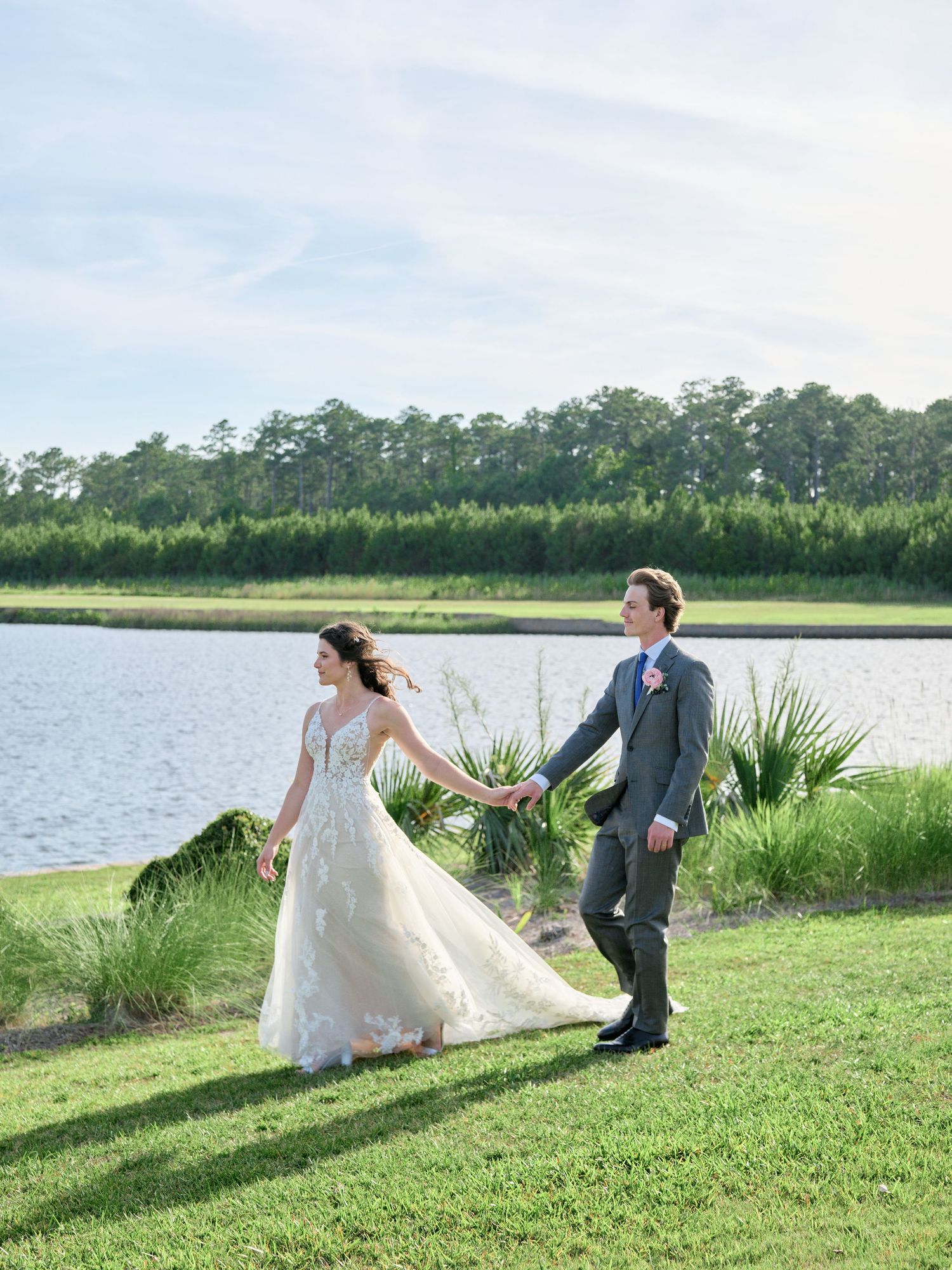 A couple in wedding attire holds hands while walking on a grassy lawn beside a serene waterfront with pine trees in background.