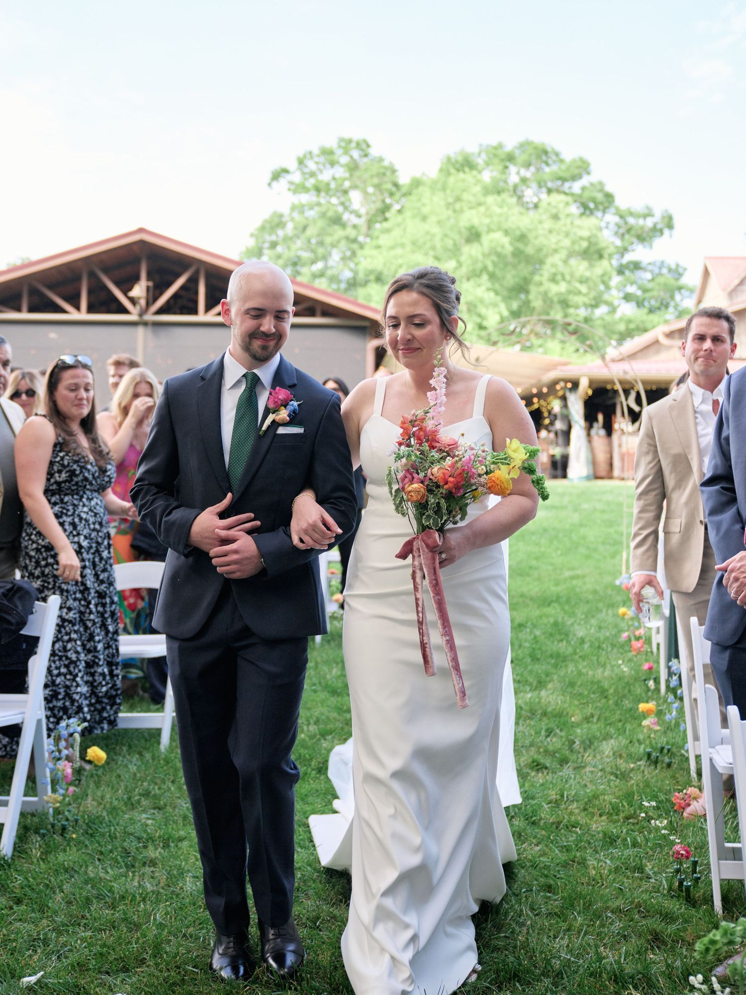 Wedding couple walks down aisle at outdoor venue with rustic building in background.