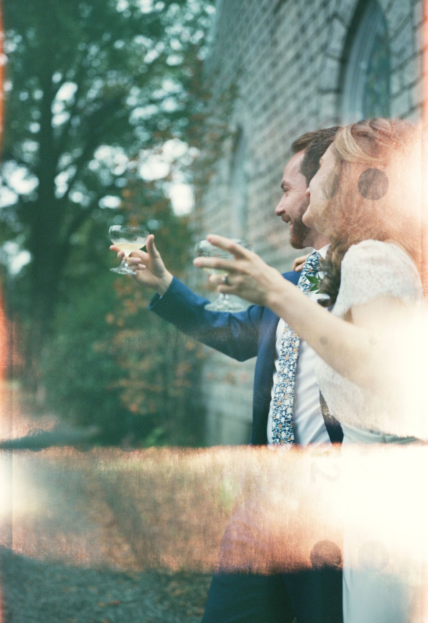 Wedding guests raise their glasses in celebration against a sunlit brick wall backdrop.